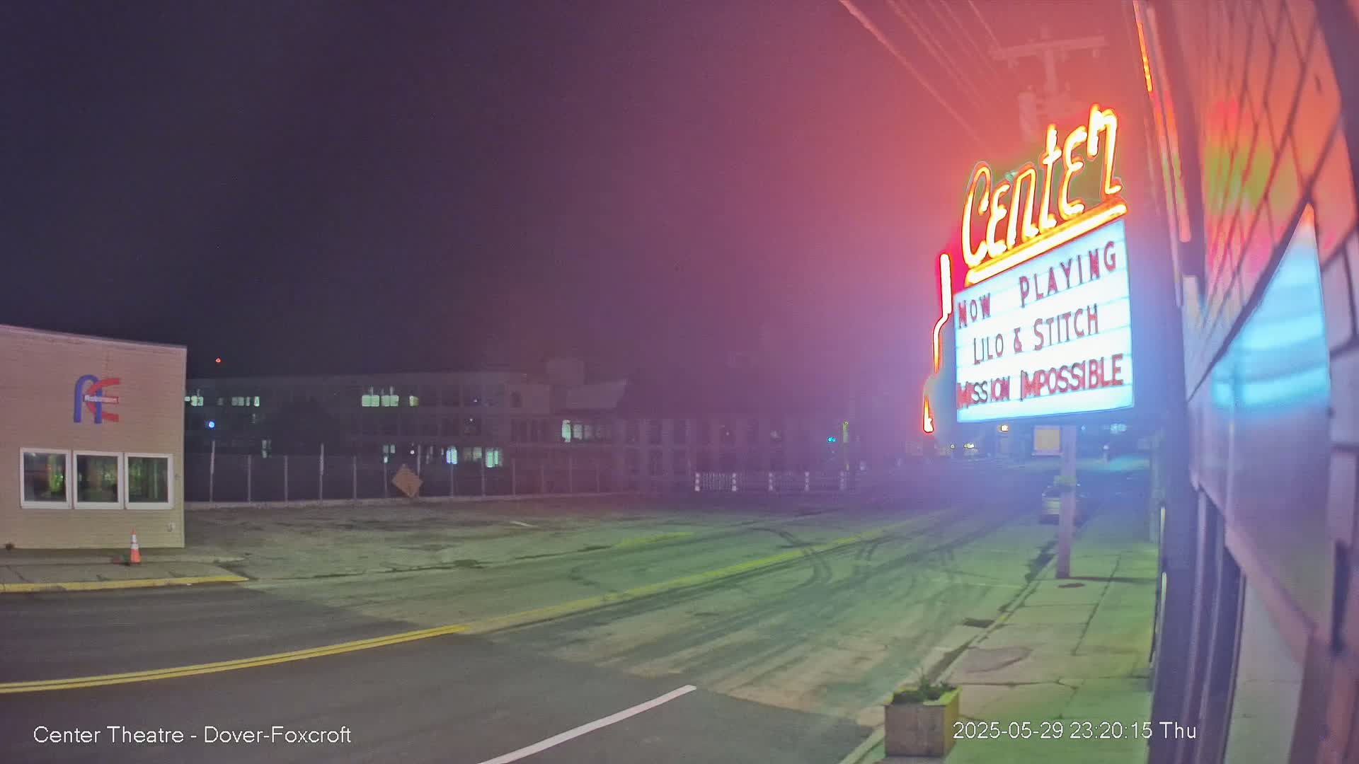 A nighttime view of a mostly empty parking lot and street in front of a movie theater marquee advertising "Lilo & Stitch" and "Mission Impossible," under a dark, slightly hazy sky.