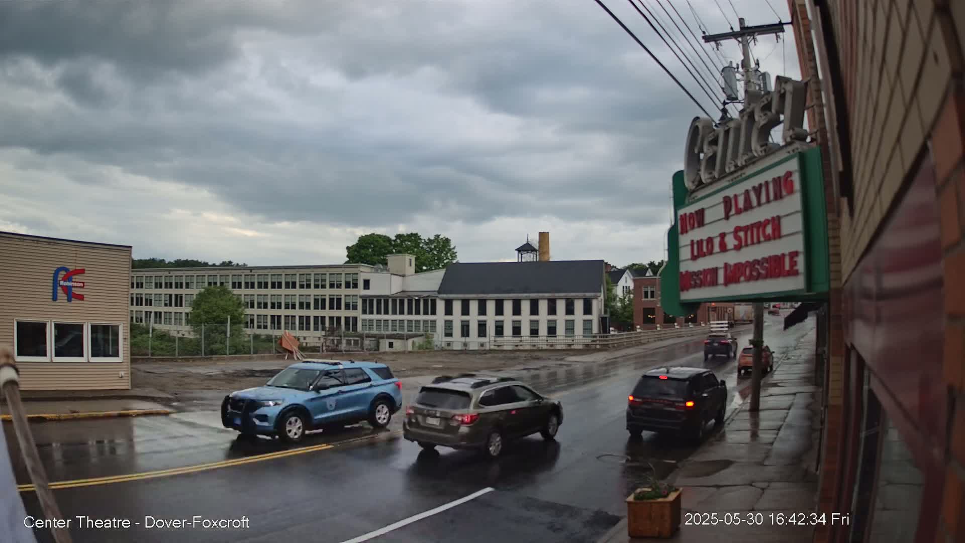 Several cars drive on a rain-slicked street in front of a theater and older factory buildings under a cloudy sky.