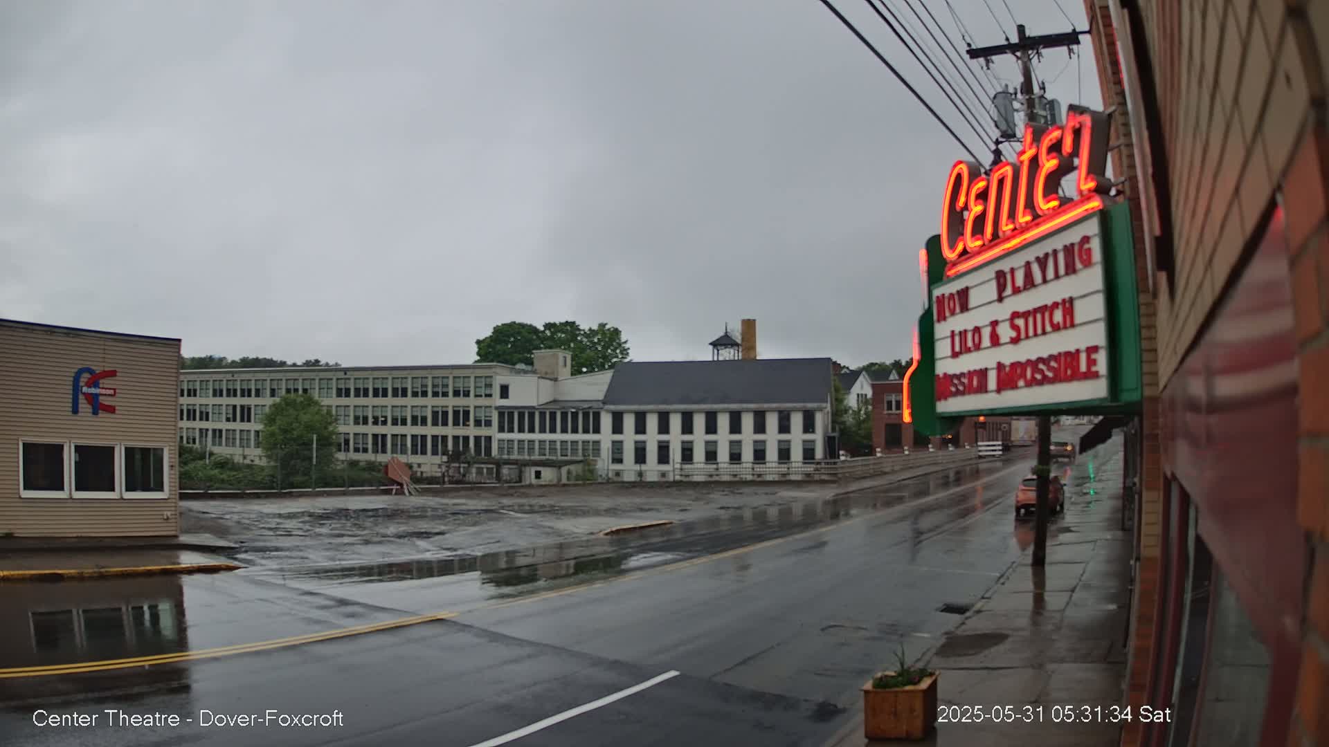 A wet street scene on a cloudy day shows a movie theater marquee, several buildings, and a mostly empty parking lot.
