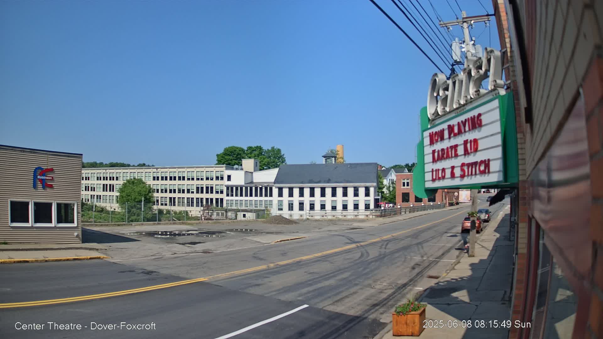 A sunny day reveals a street scene with several buildings, including a long factory and a smaller, older building, with a relatively empty street and a marquee showing movie titles.