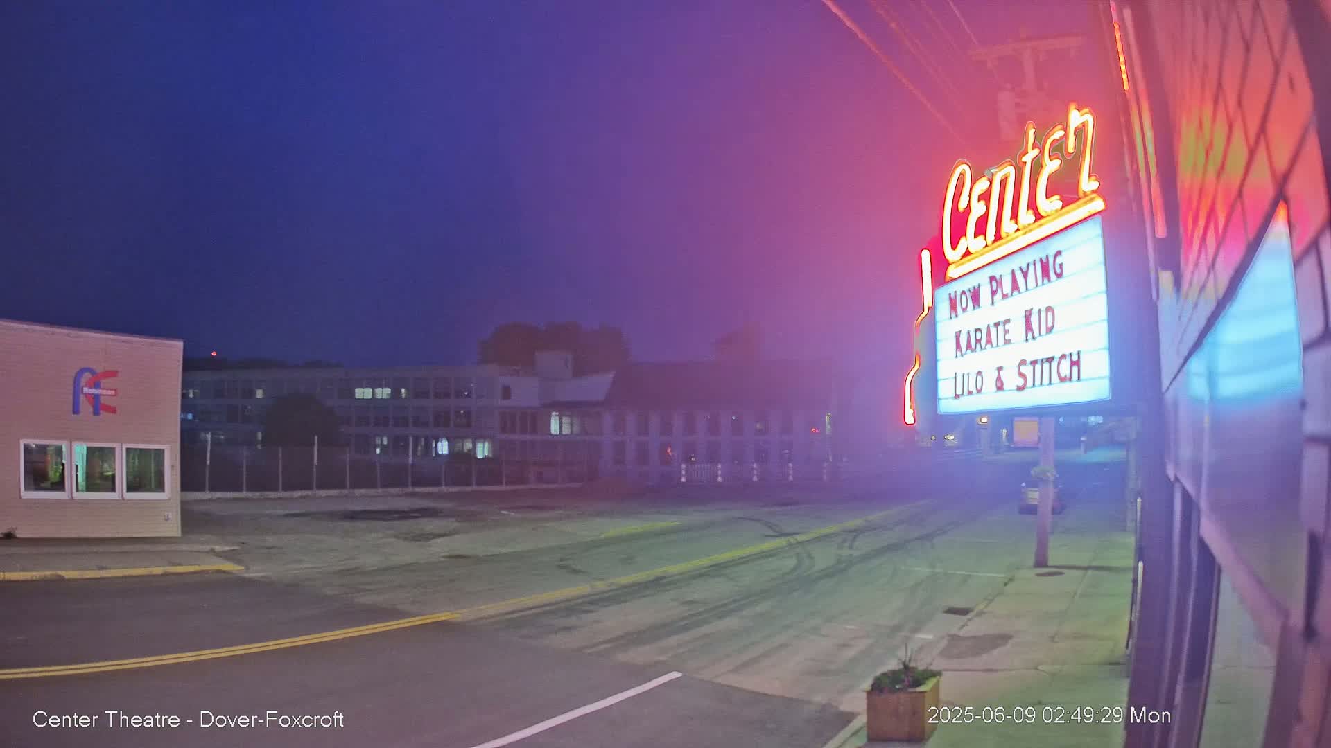 A mostly empty street at night, under a dusky sky, shows a movie theater marquee advertising "Karate Kid" and "Lilo & Stitch".