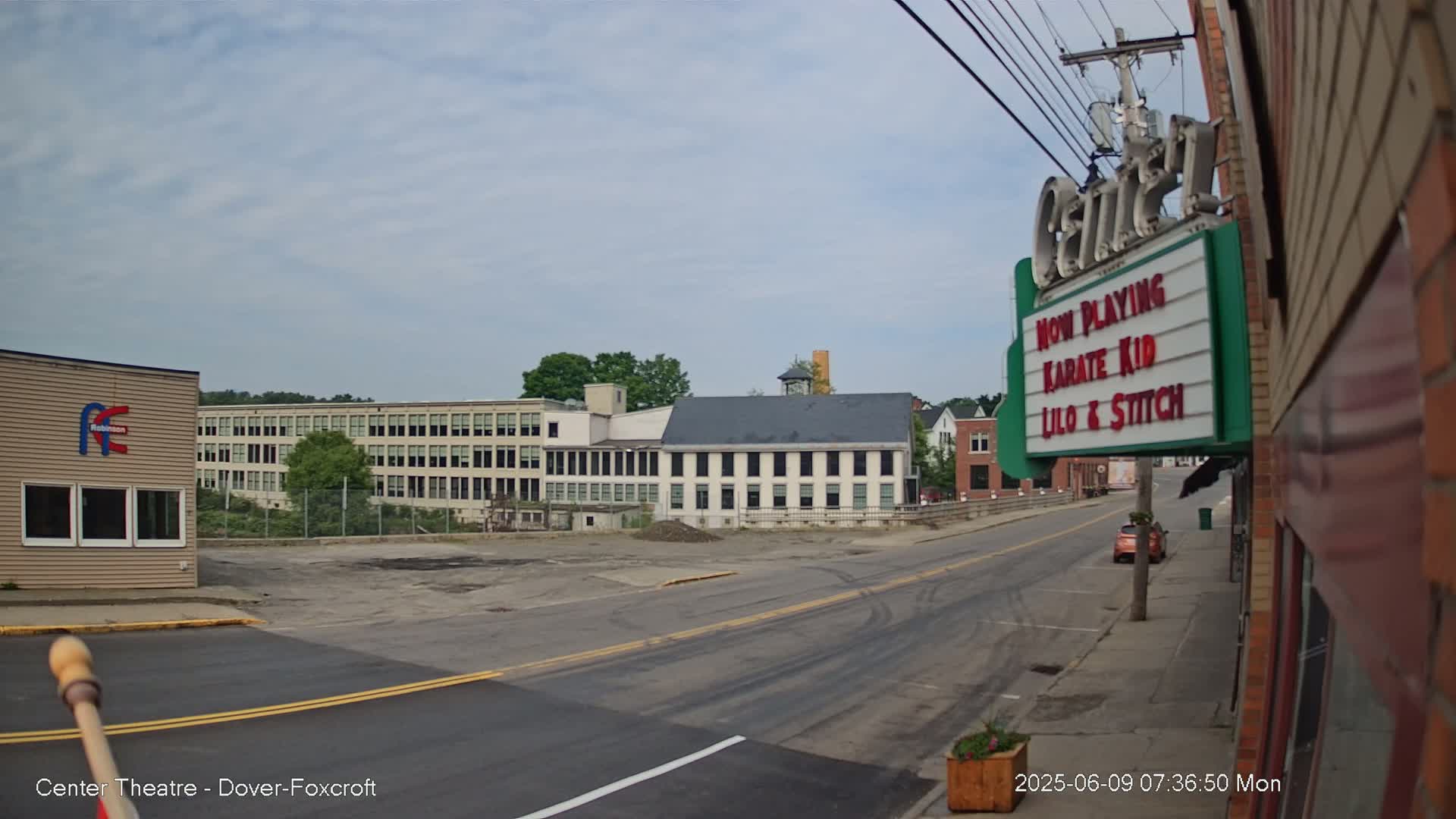 A mostly sunny day reveals a street scene with several buildings, including a large factory, a smaller multi-story building, and a movie theater marquee advertising "Karate Kid" and "Lilo & Stitch."