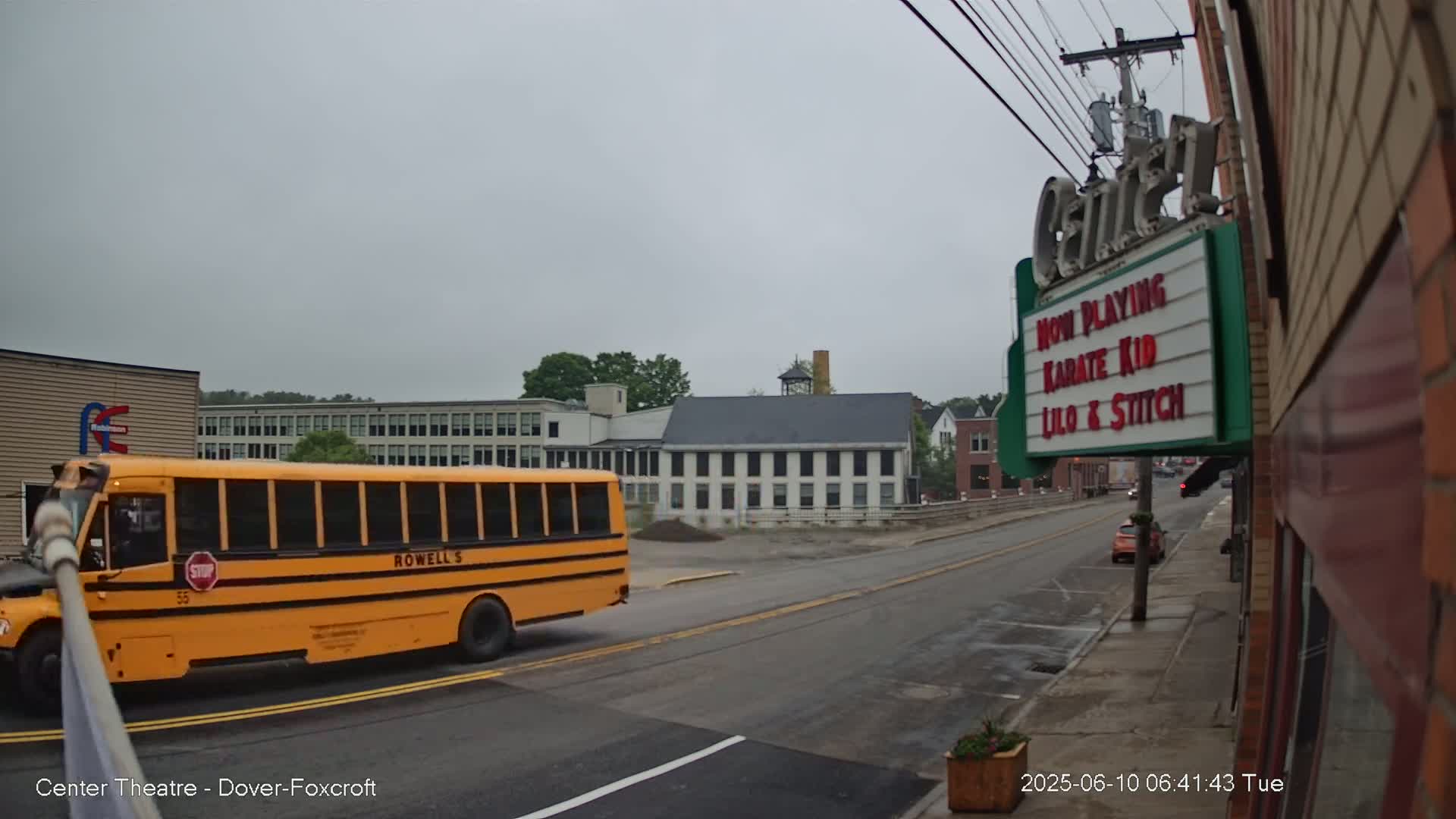 A yellow school bus drives past a street with overcast skies,  a movie theater marquee, and several buildings including a large, white factory-like structure.