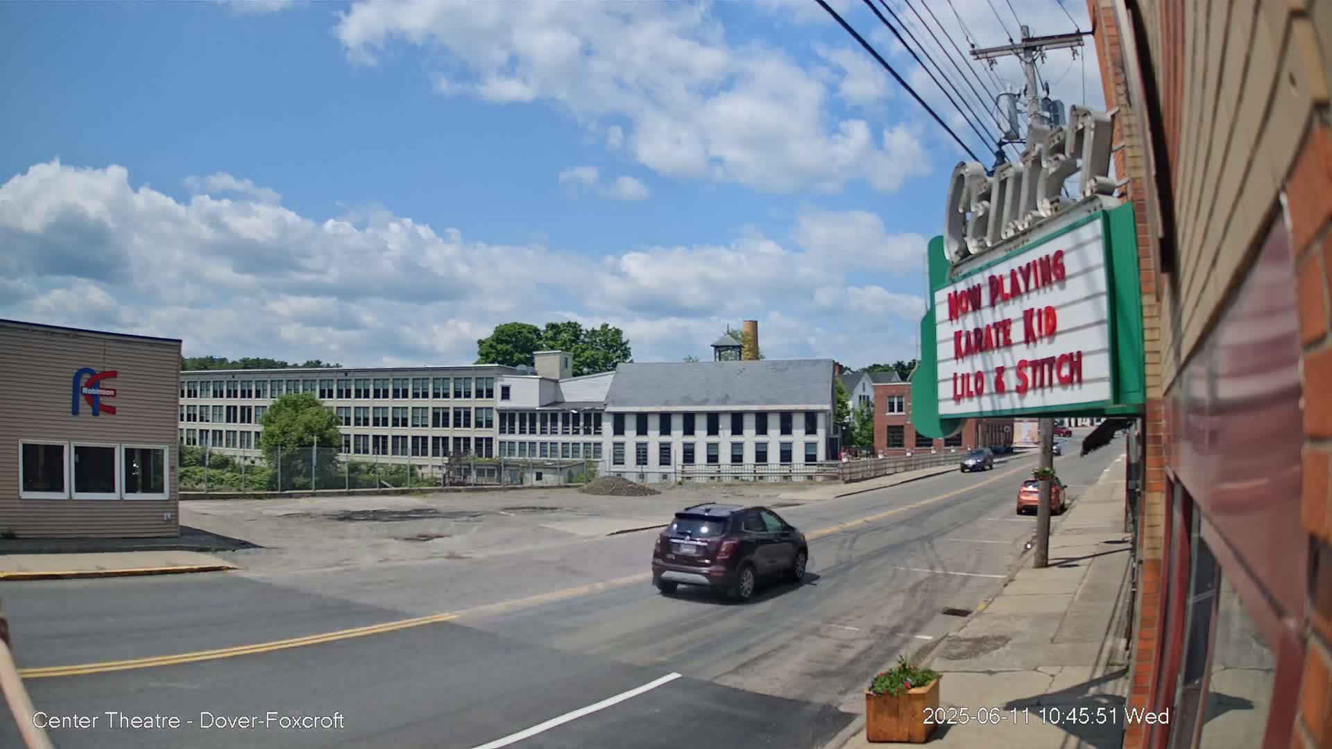 A partly cloudy day reveals a street scene with a car driving past a movie theater marquee advertising "Karate Kid" and "Lilo & Stitch," and a large industrial building in the background.