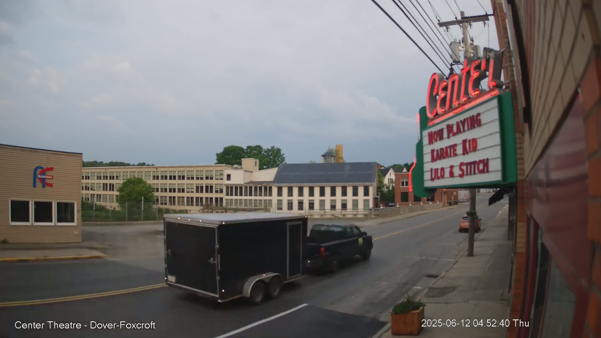 A black enclosed trailer is being towed down a street past a movie theater marquee under an overcast sky.