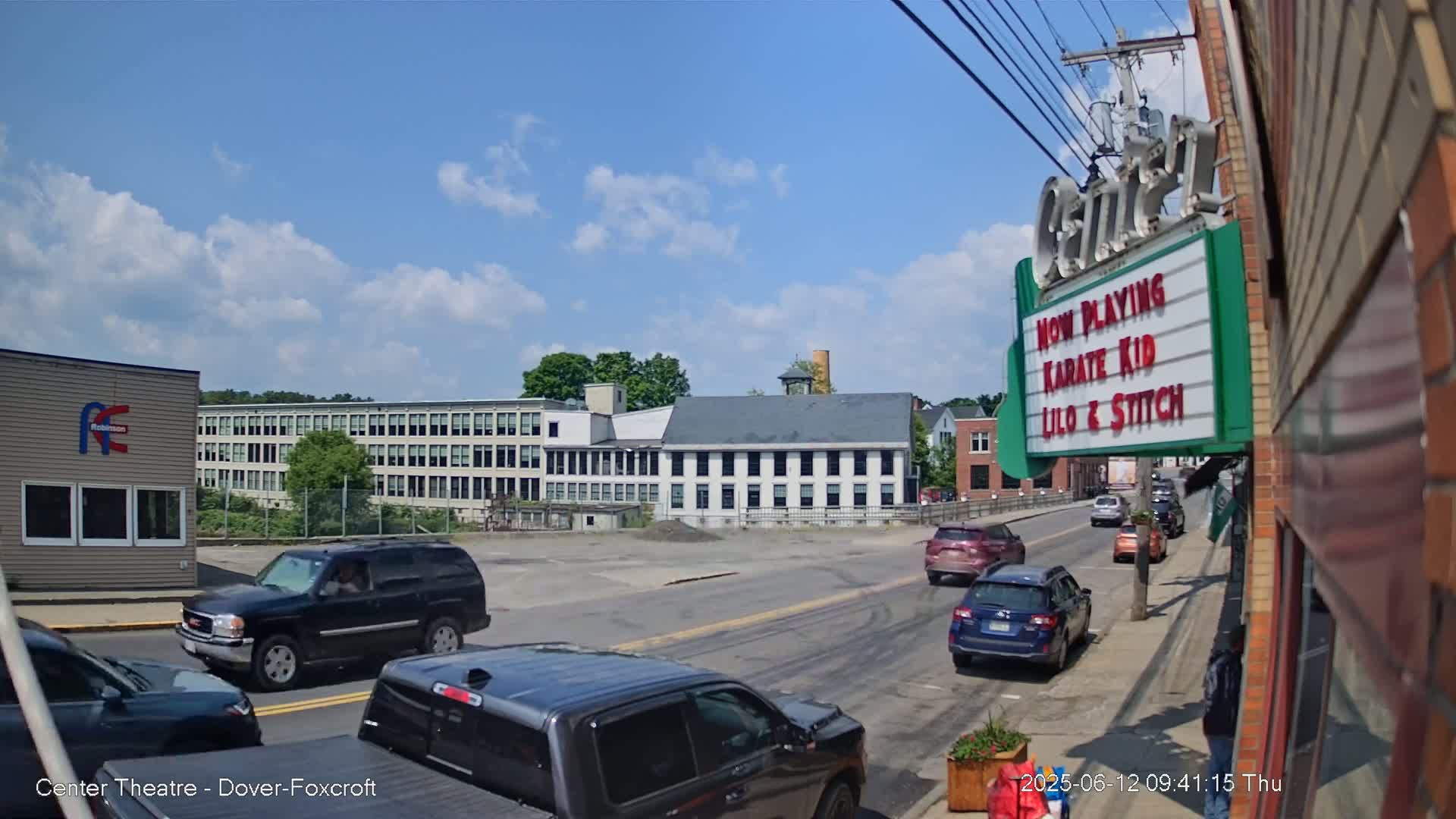 Under a partly cloudy blue sky, a street scene shows several cars driving past a large industrial building and a smaller, older building, with a movie theater marquee in the foreground advertising "Karate Kid" and "Lilo & Stitch."