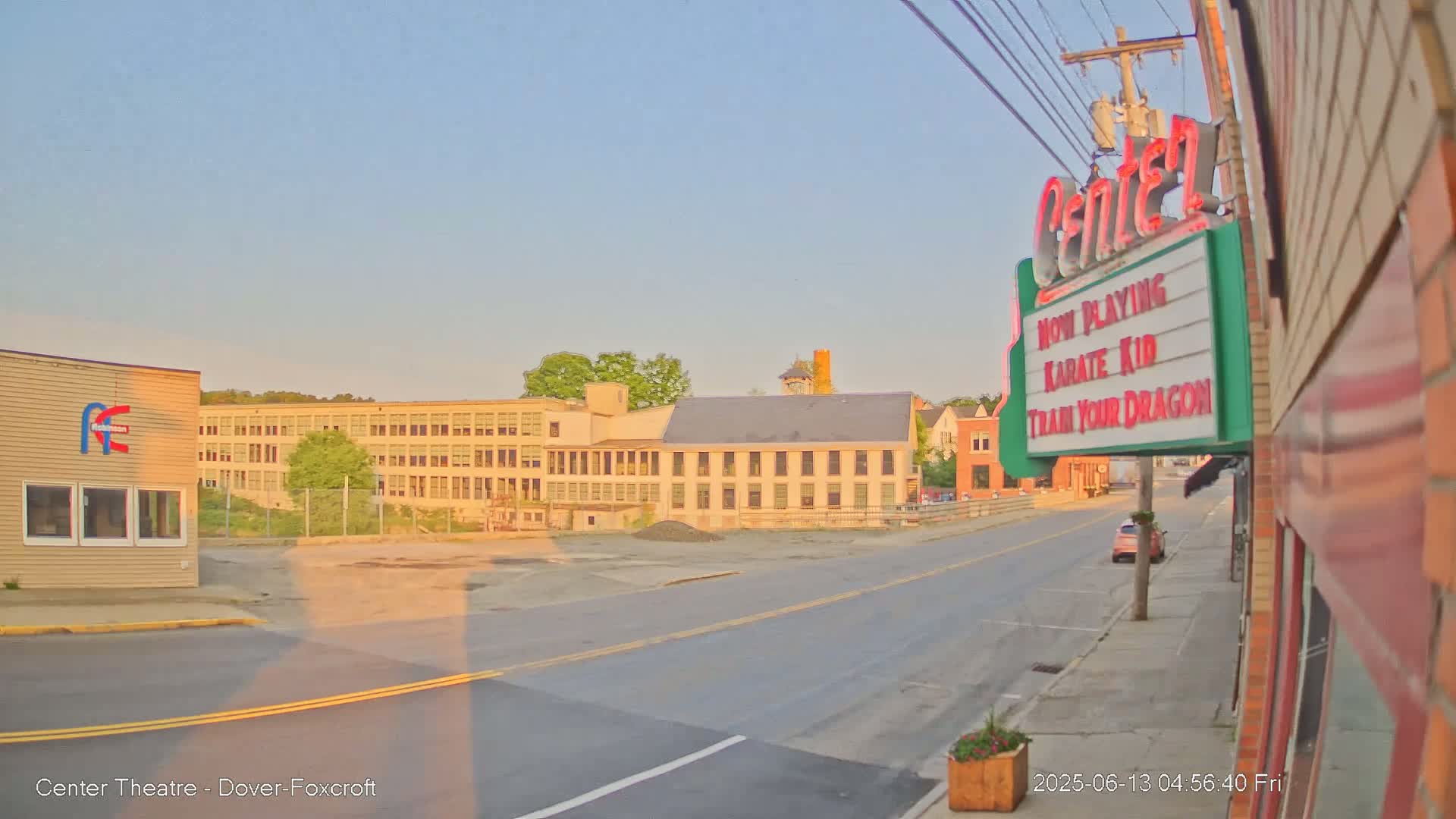 A long, empty road stretches toward a large light-beige building under a clear, light-blue sky at sunrise.