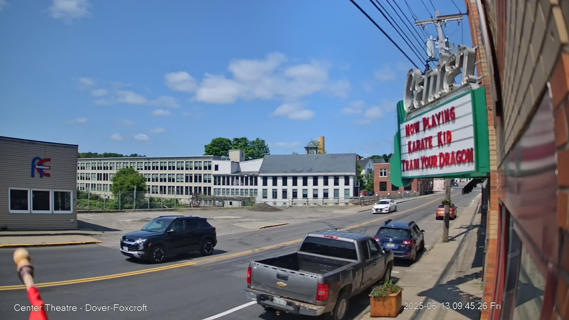 A sunny day shows several vehicles on a street in front of industrial buildings and a movie theater marquee advertising "Karate Kid" and "Train Your Dragon."