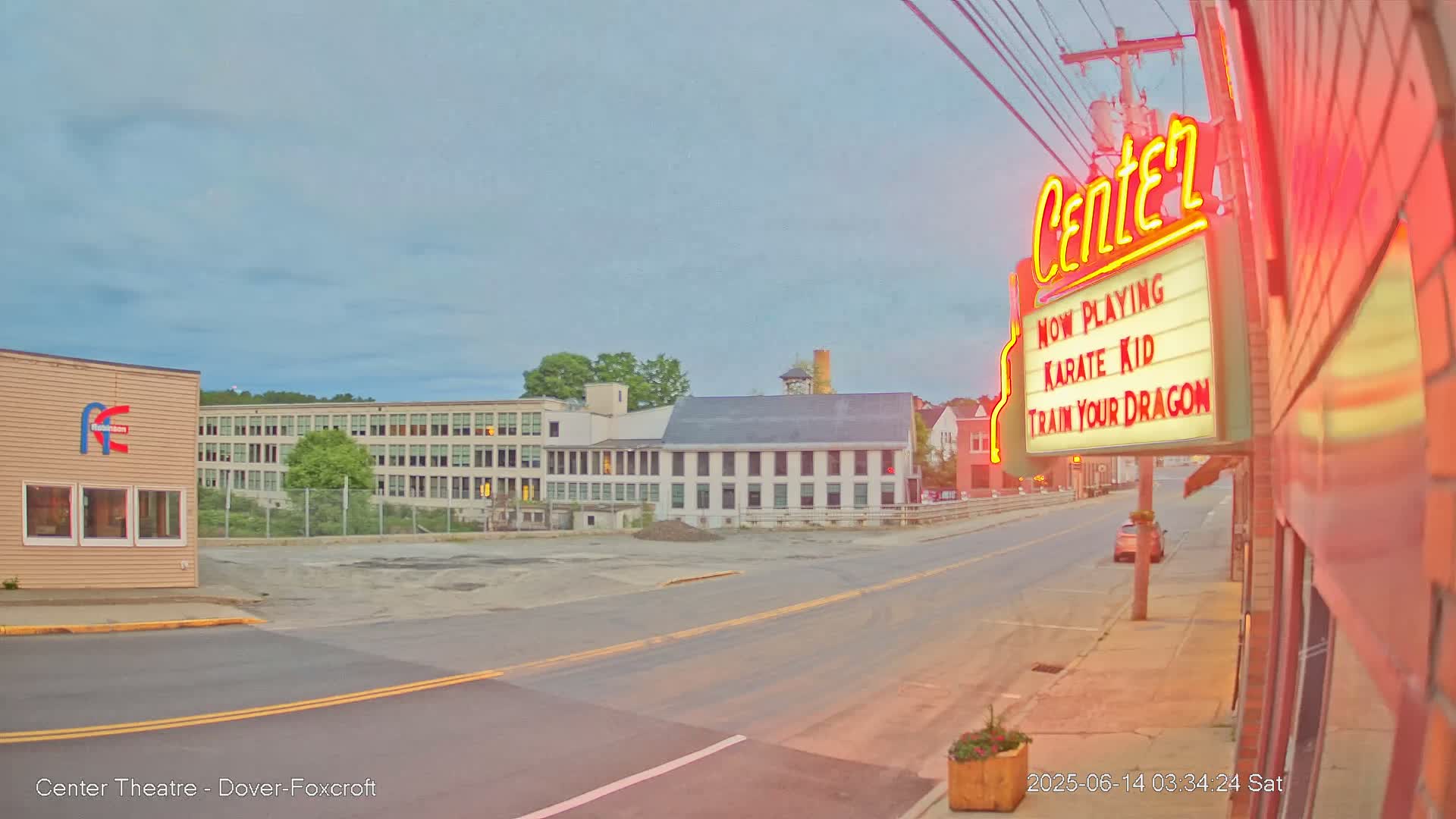 A street scene at dusk shows a movie theater marquee advertising "Karate Kid" and "Train Your Dragon," across the street from several large buildings, including a multi-story factory-like structure.