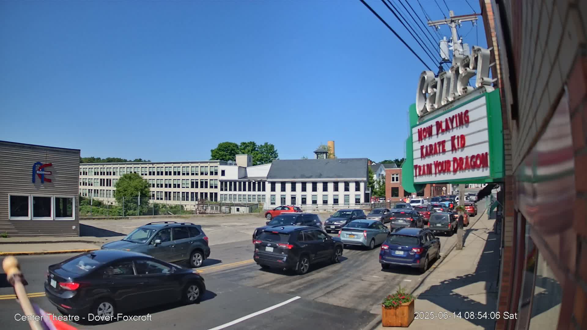 A sunny day reveals a street scene with a line of cars stopped at an intersection in front of a movie theater marquee, and several industrial-style buildings in the background.