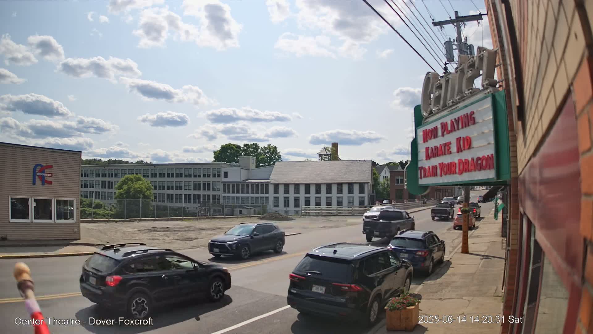 A sunny day reveals a street scene with a line of cars stopped at an intersection in front of a movie theater marquee, and several industrial-style buildings in the background.