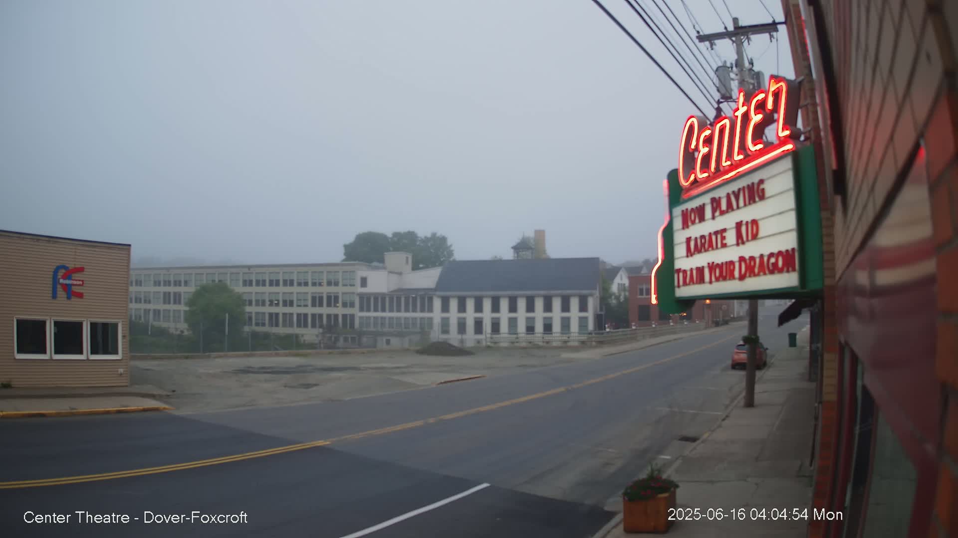 A foggy street scene shows a road leading toward several buildings, including a large factory and a smaller building with a movie marquee.