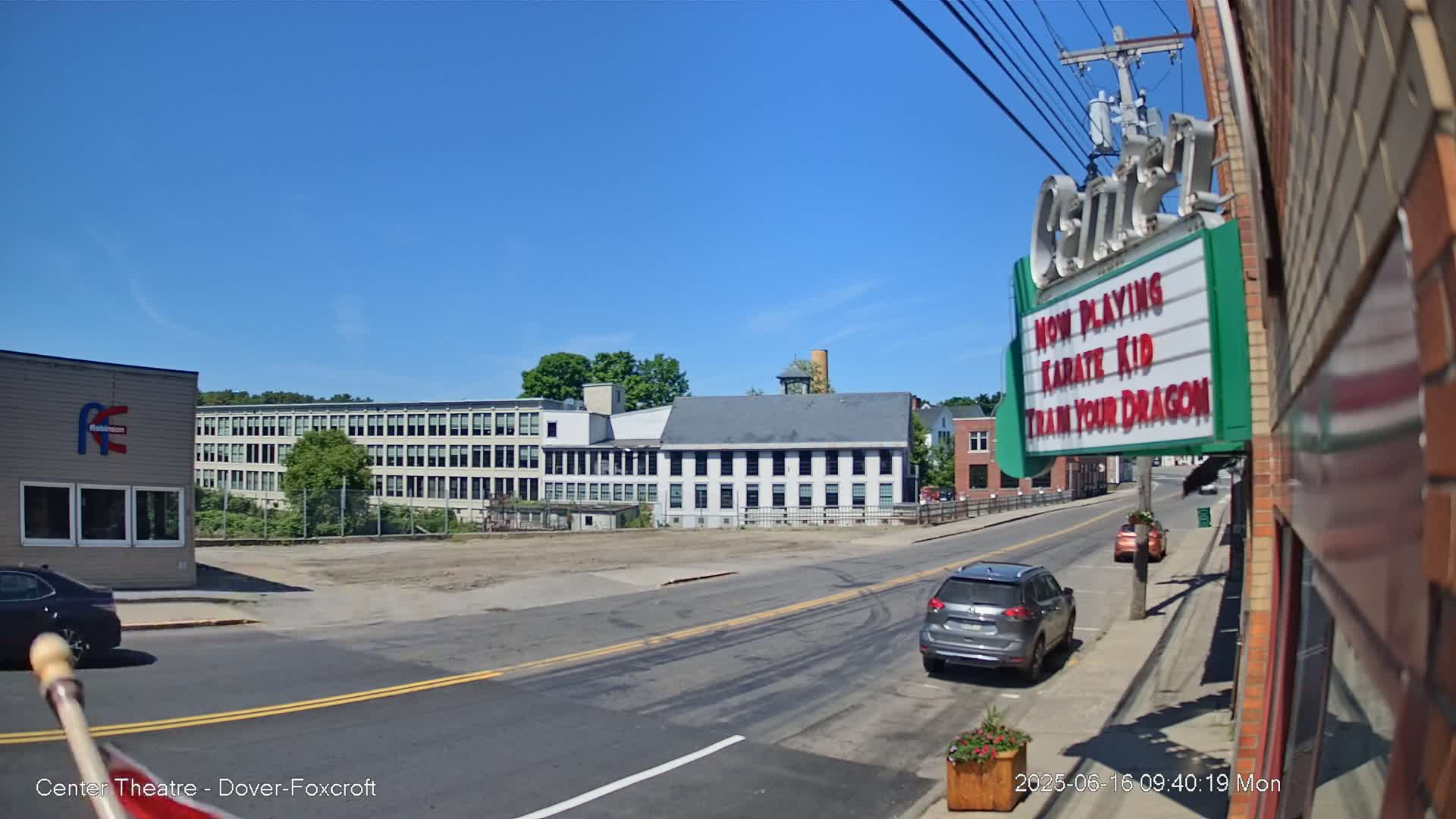 A sunny day reveals a street scene with several buildings, including a large multi-story factory, smaller commercial buildings, and a movie theater marquee advertising "Karate Kid" and "Train Your Dragon."