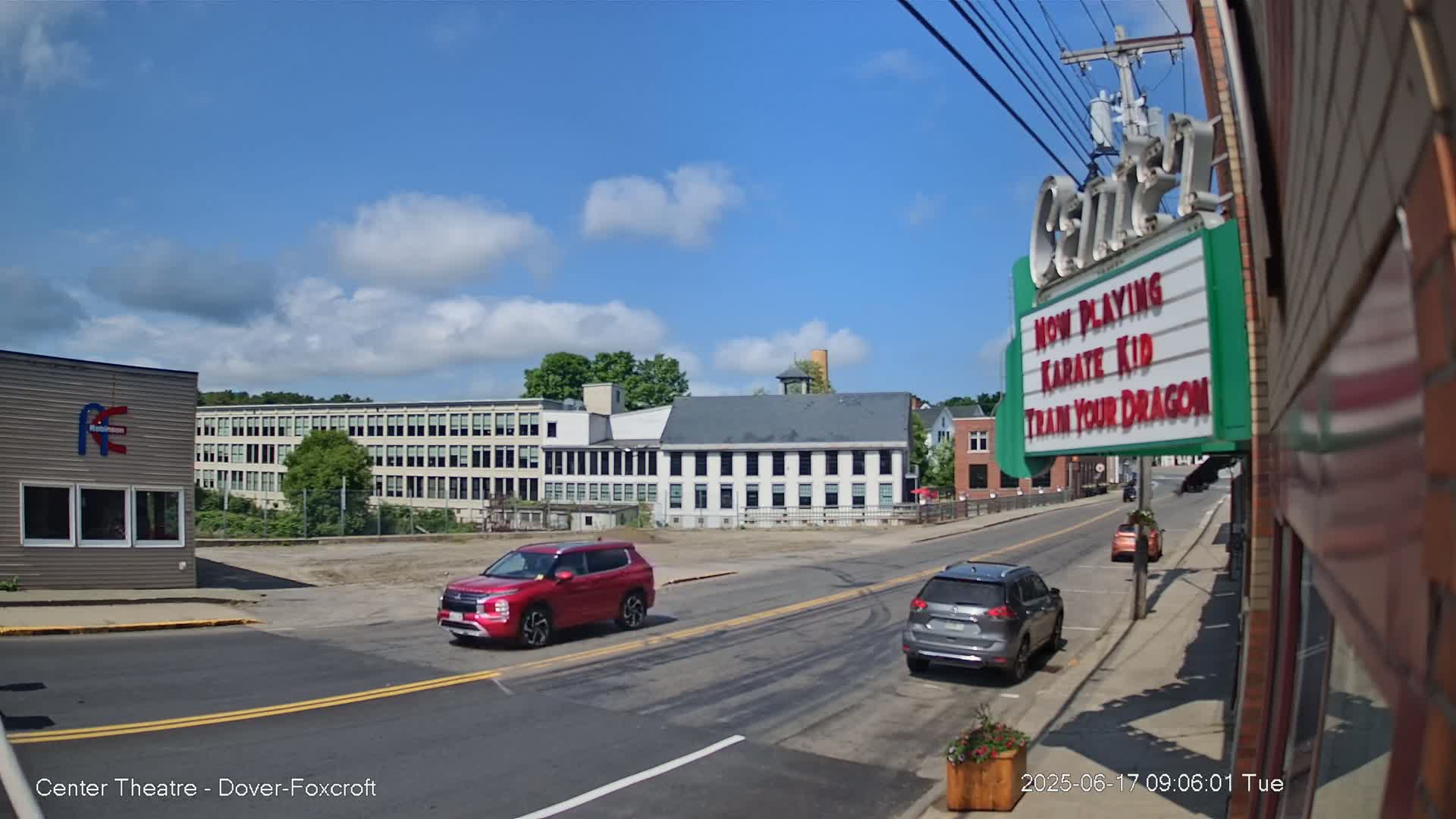 A sunny day reveals a street scene with two SUVs and several buildings, including a large multi-story factory-like structure and a smaller, older building across the street from a movie theater marquee advertising "Karate Kid" and "Train Your Dragon."