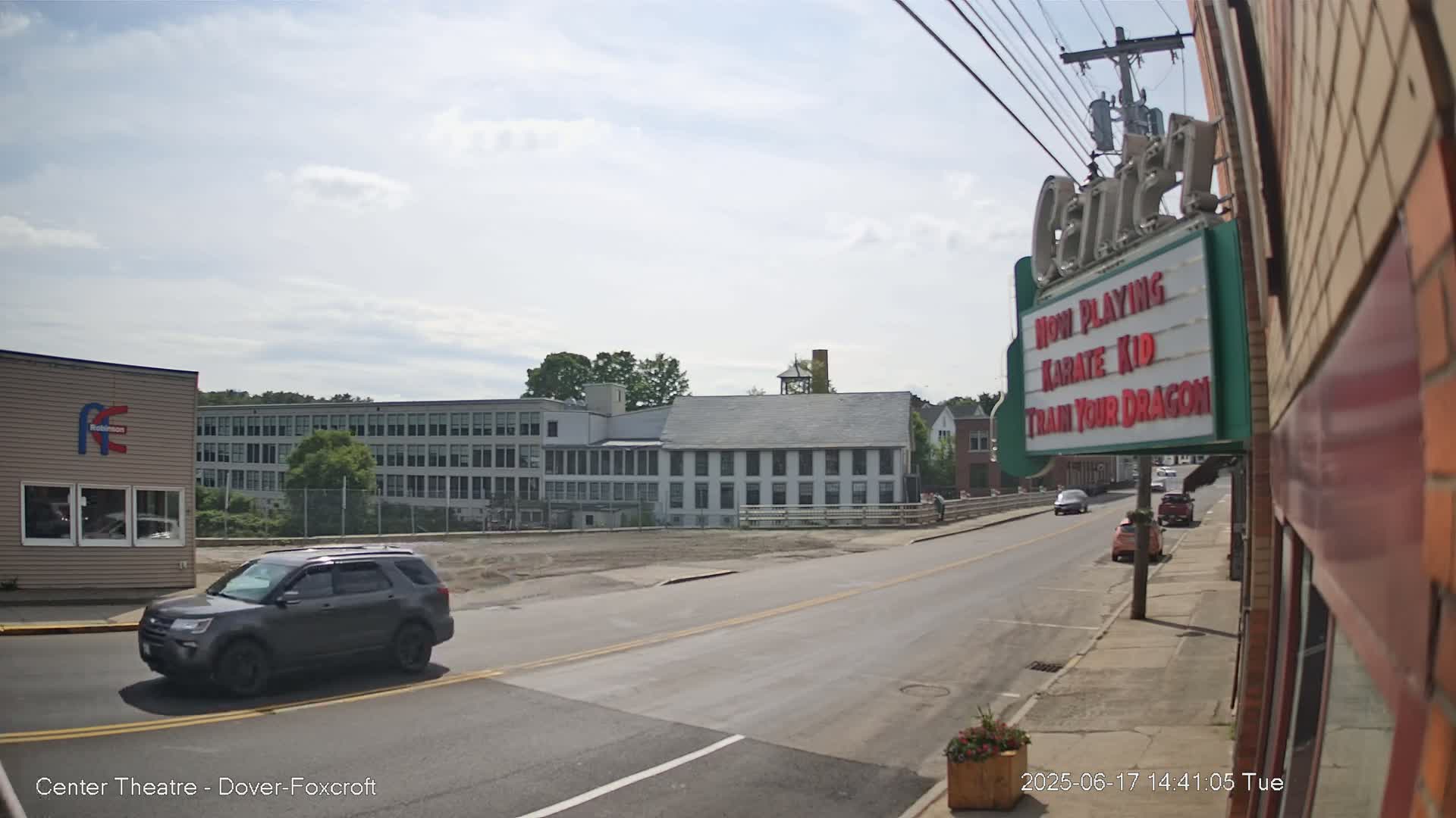 A gray SUV drives past a street scene on a partly cloudy day, with a large building in the background and a movie theater marquee advertising "Karate Kid" and "Train Your Dragon."
