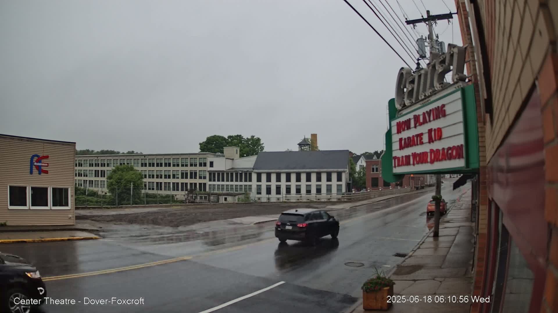 A wet street scene shows a dark-colored SUV driving past a large, light-colored building under a gray, overcast sky.