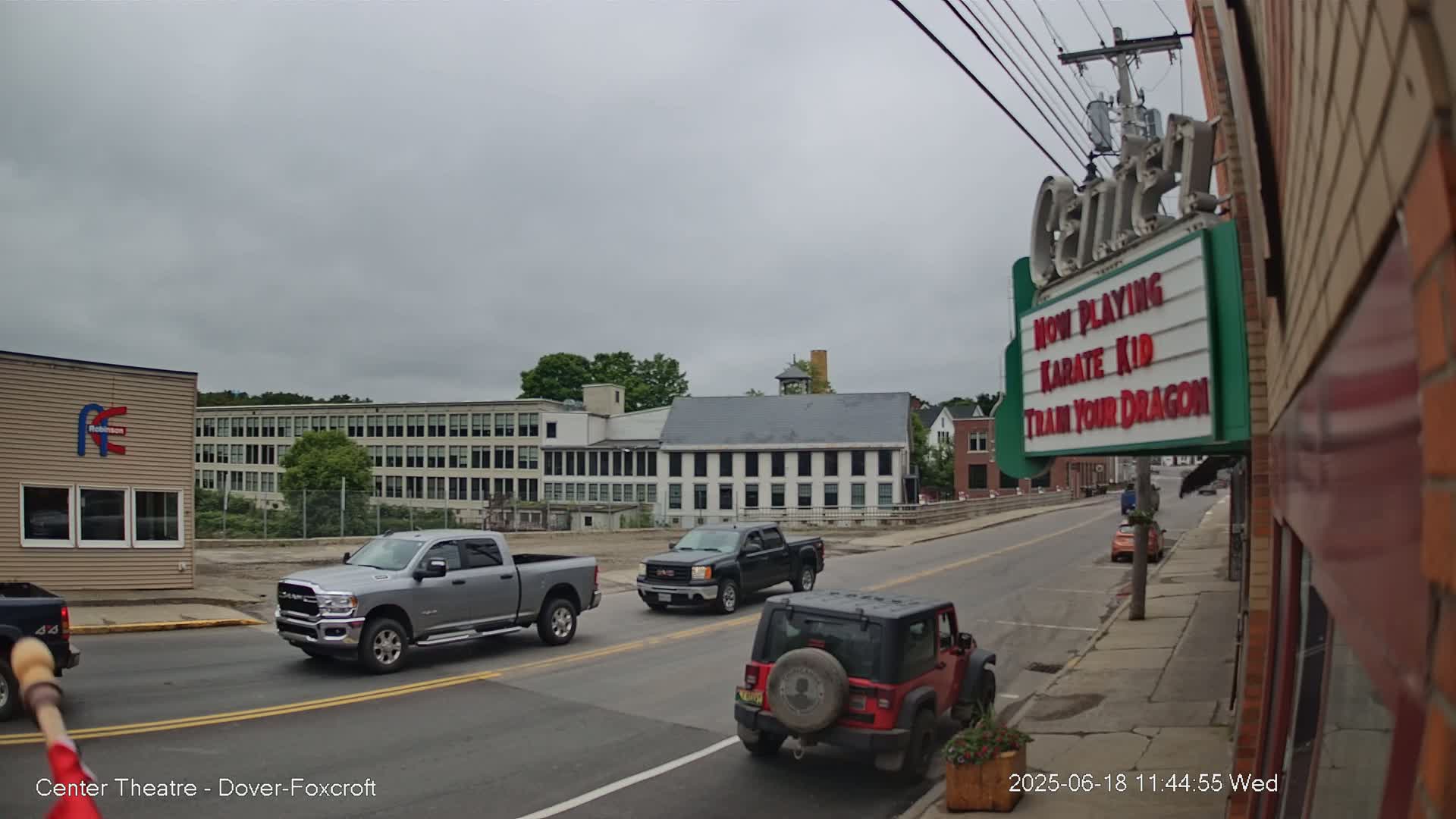A street scene on an overcast day shows several vehicles at an intersection near a large building and a movie theater marquee advertising "Karate Kid" and "Train Your Dragon".