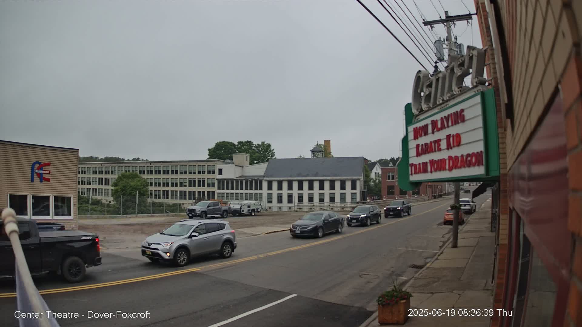 Several vehicles are driving on a street in front of a multi-story building under an overcast sky.