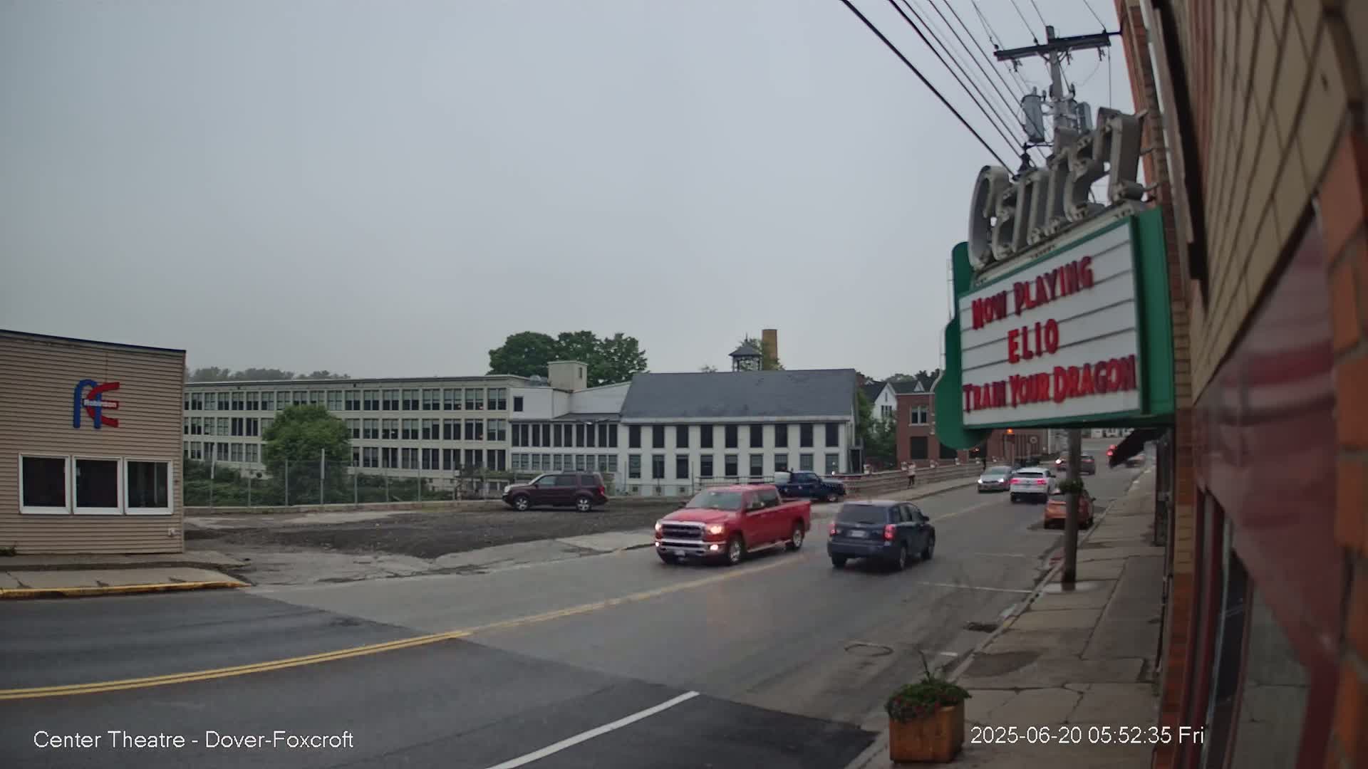 Several vehicles drive on a street in front of buildings under an overcast sky.