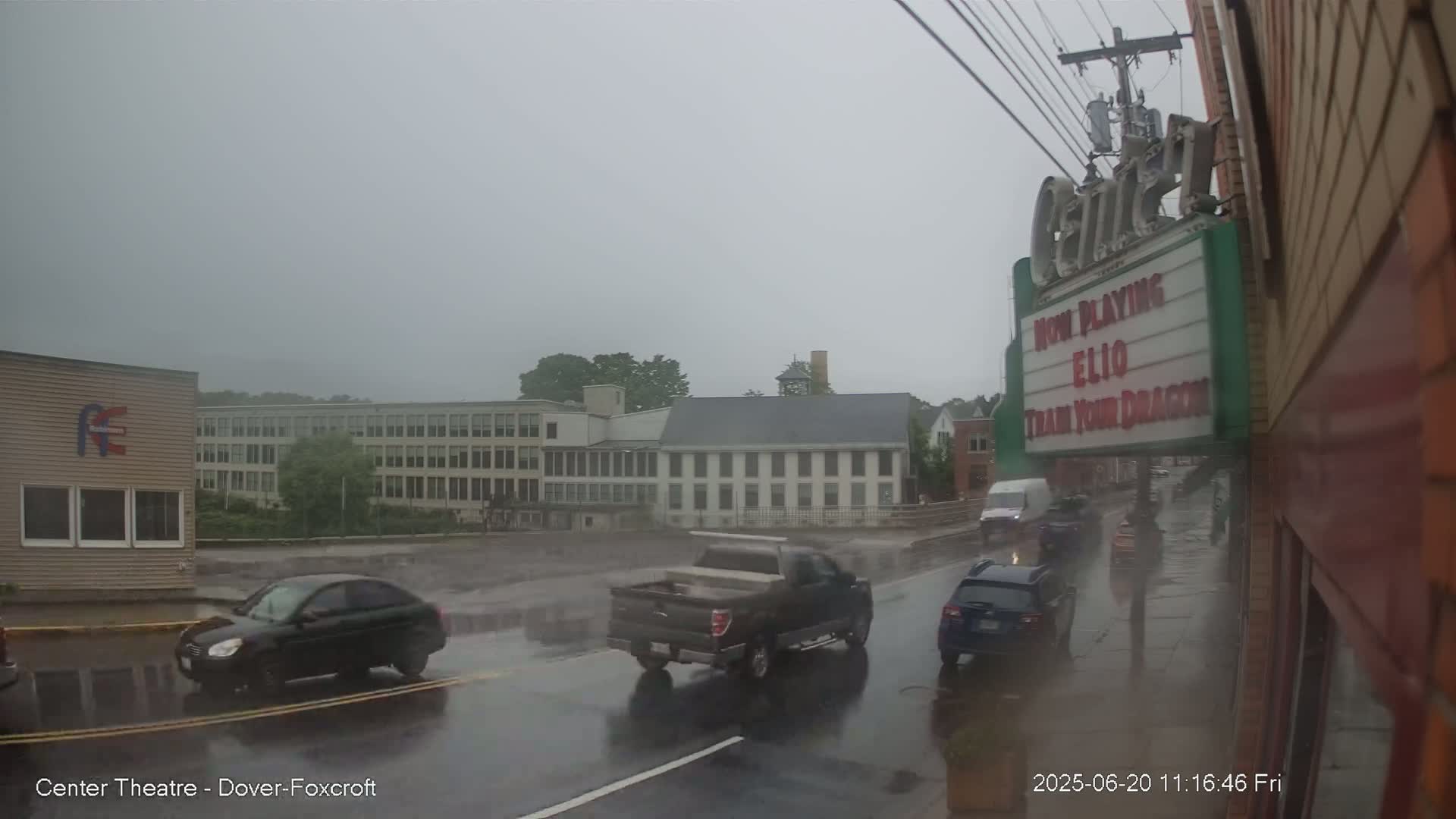 A rainy street scene shows several vehicles driving past buildings, including a large multi-story structure and a theater marquee.