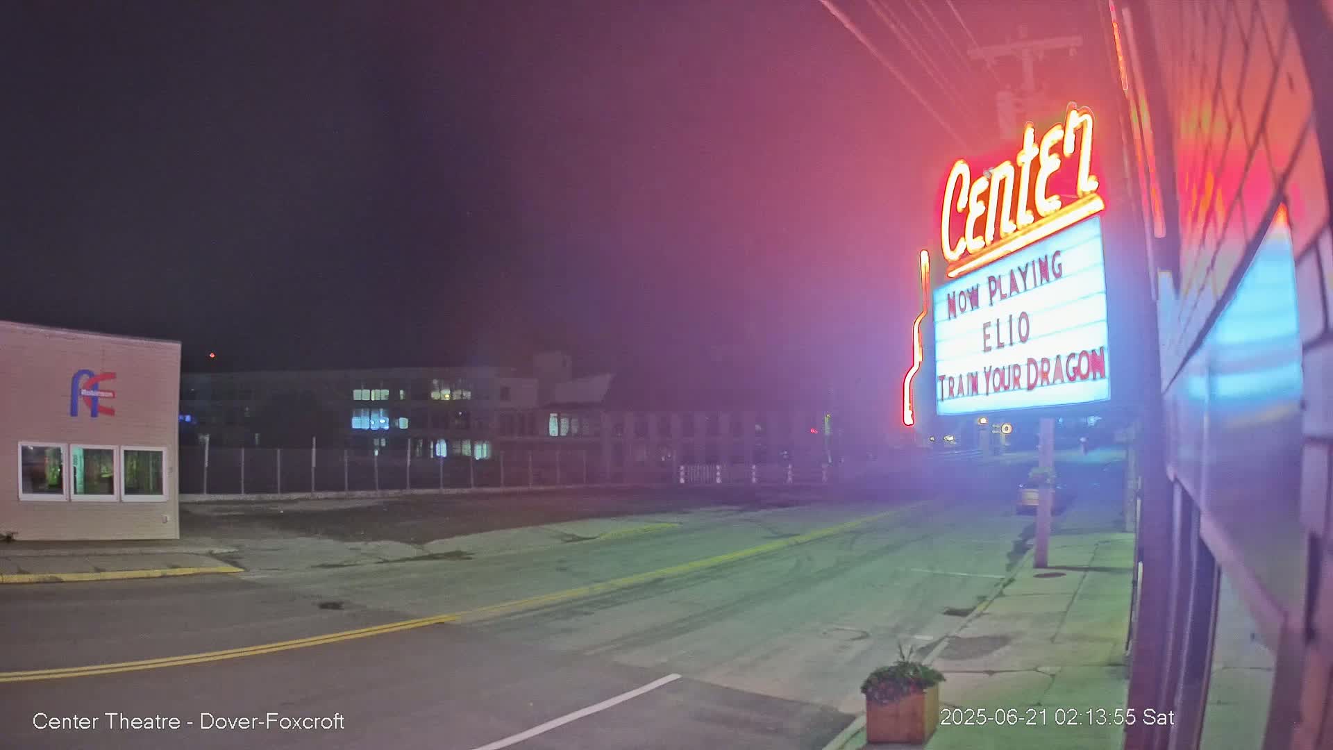 A nighttime street scene shows a movie theater marquee advertising "Elio" and "Train Your Dragon," with a building and empty parking lot visible under a hazy, dark sky.