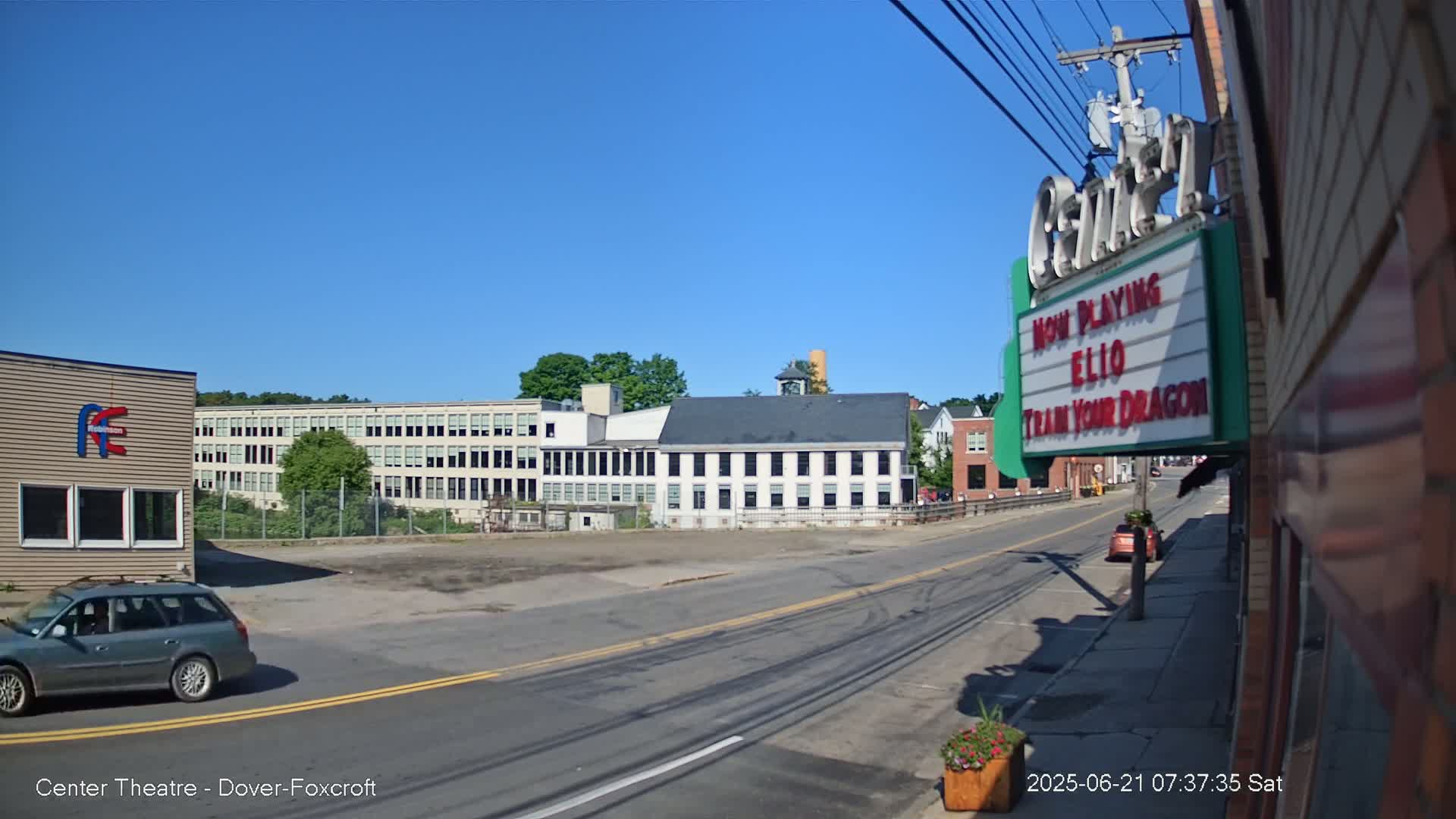 A sunny day reveals a street scene with a multi-story building, a large factory-like structure, and a movie theater marquee advertising "Ellio" and "Train Your Dragon."