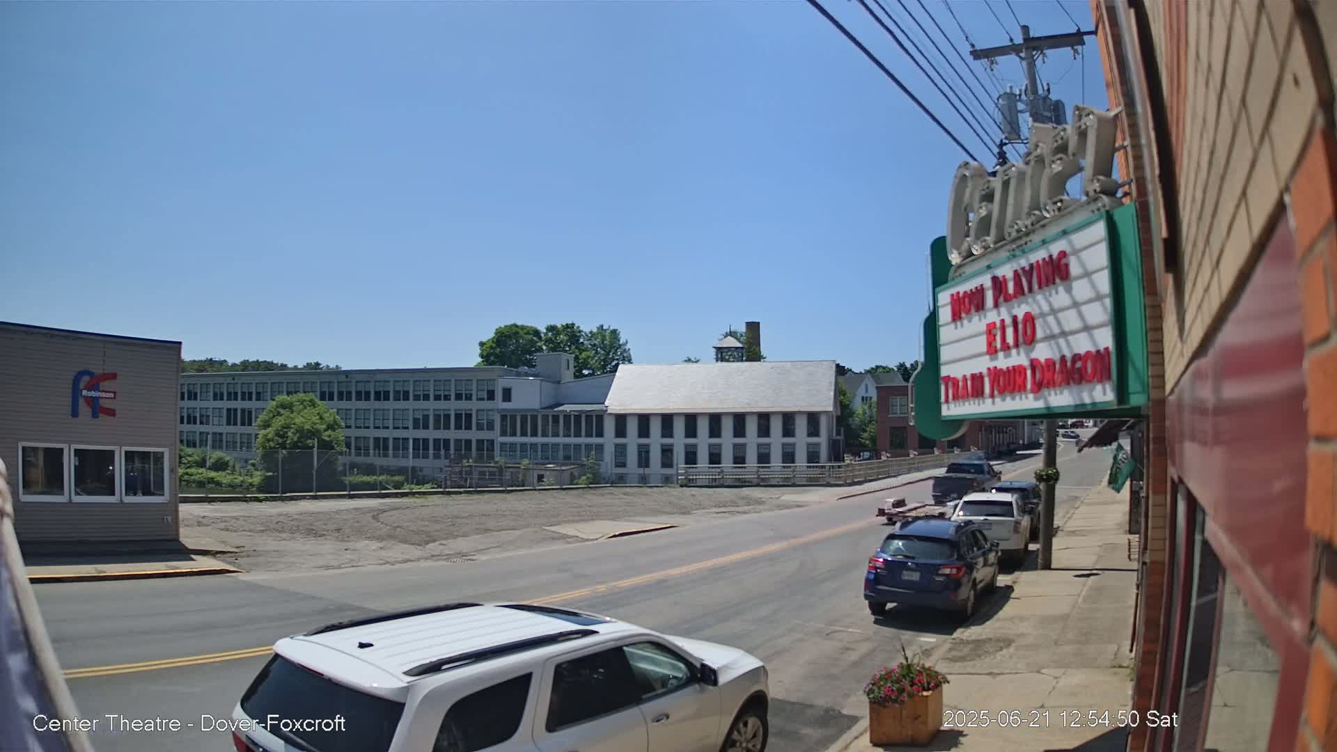 A sunny day reveals a street scene with parked cars, a multi-story building in the background, and a movie theater marquee.