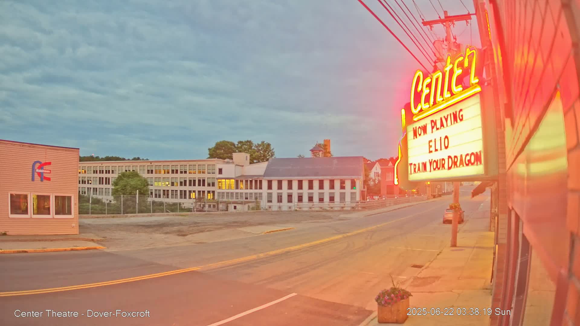 A street scene under a mostly cloudy, twilight sky shows a movie theater marquee and several buildings, including a large multi-story structure.
