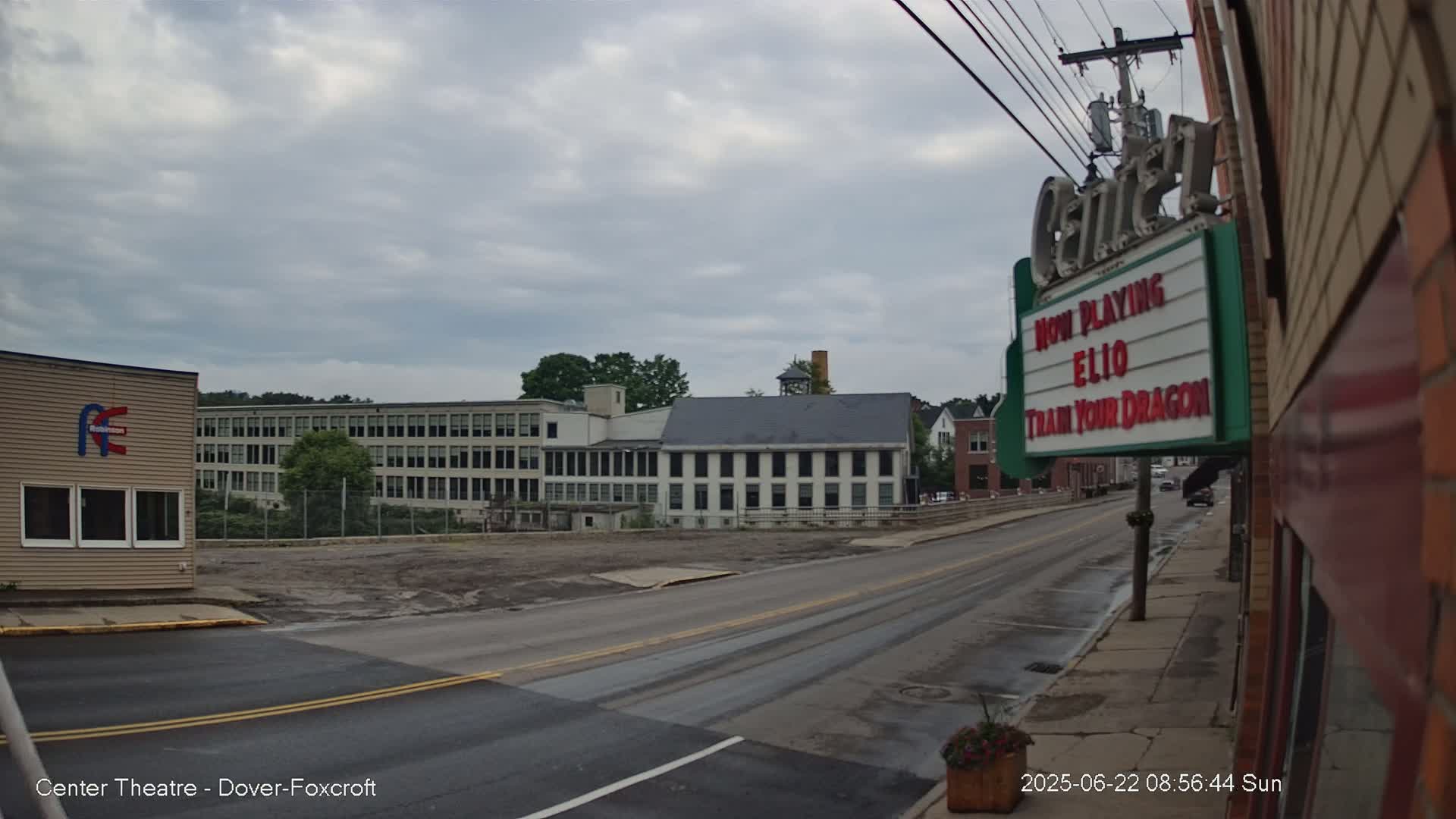 A street scene under a cloudy sky shows a long, light-colored multi-story building across the street from a movie theater marquee, with other buildings and a mostly empty lot visible in the background.