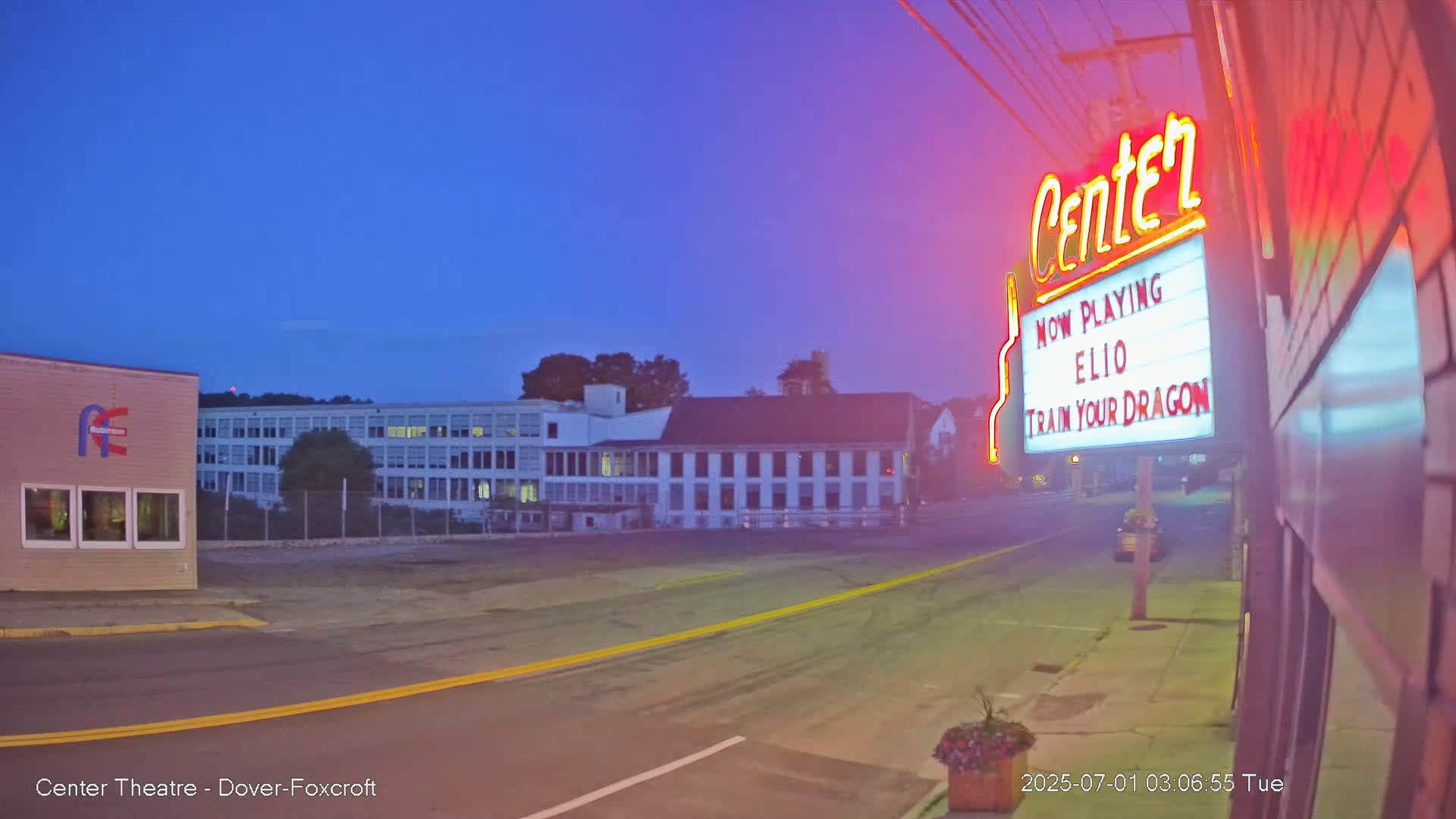 A mostly empty street at twilight, with a movie theater marquee displaying "Elio" and "Train Your Dragon," and buildings in the background.
