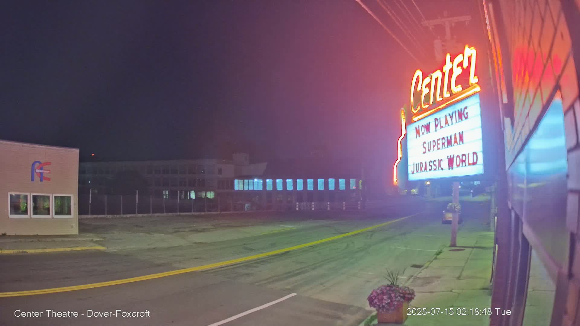 A nighttime street scene shows a movie theater marquee advertising Superman and Jurassic World, with a quiet road and some buildings visible in the background under a hazy, dark sky.
