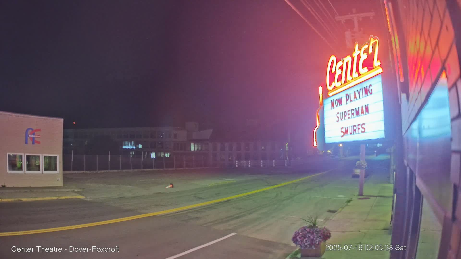 A nighttime, hazy street scene shows a movie theater marquee advertising "Superman" and "Smurfs," with a building and empty parking lot visible in the background.