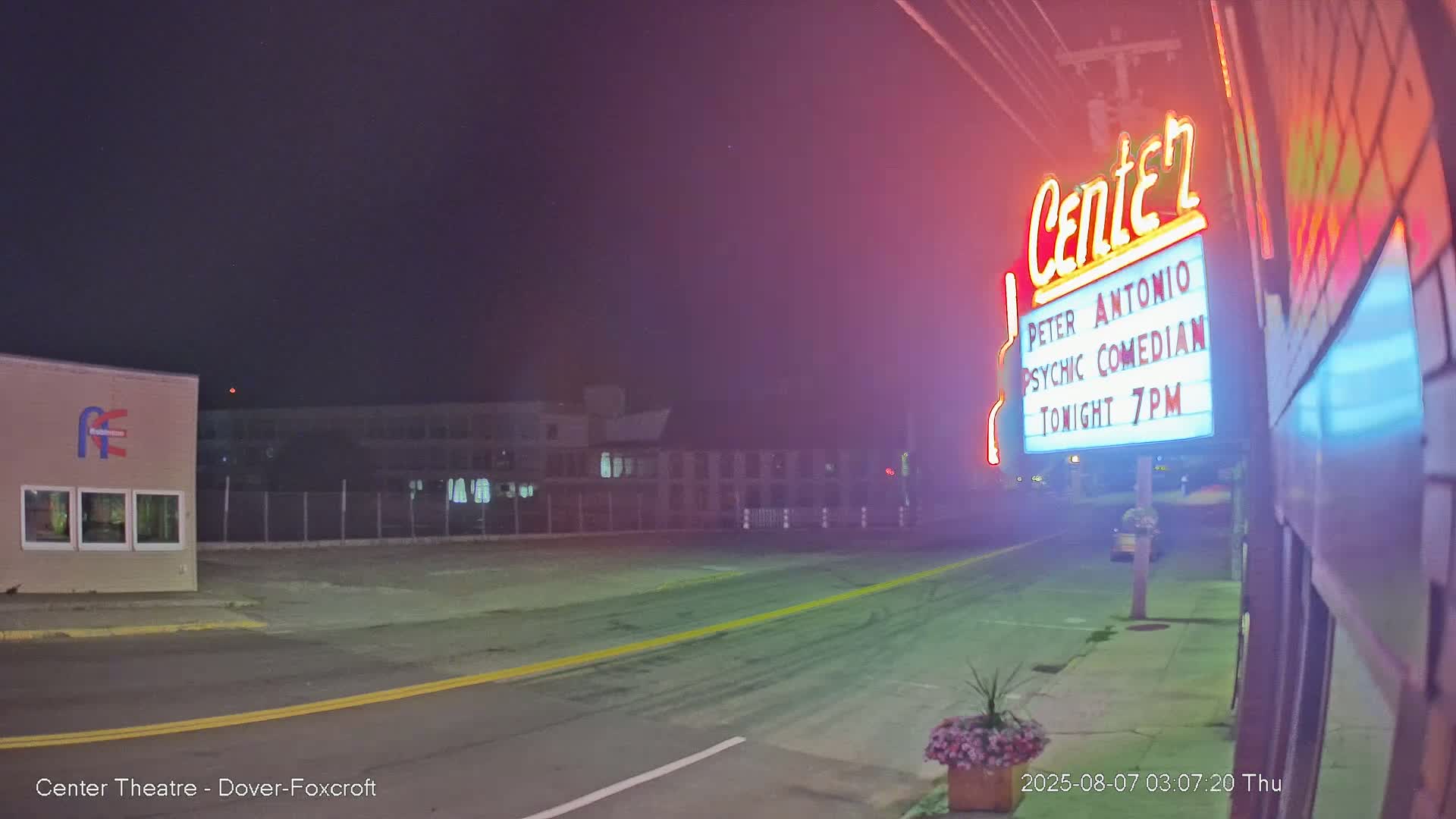 A nighttime street scene shows a brightly lit theater marquee advertising a psychic comedian, with a relatively empty road and some buildings in the background under a dark, clear sky.