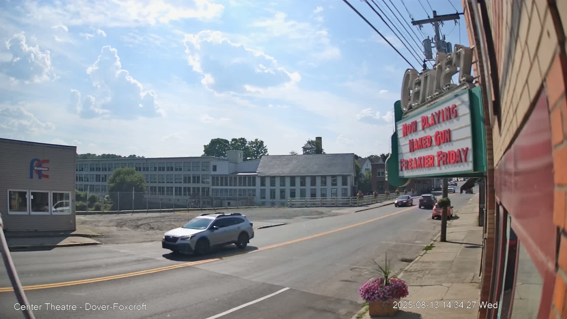 A silver SUV drives past a movie theater marquee under a partly sunny sky on a tree-lined street in front of several large buildings.