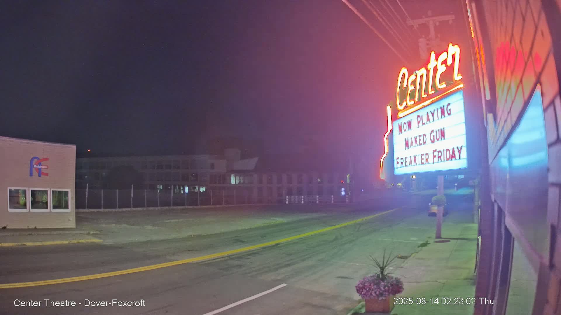 A nighttime street scene shows a movie theater marquee advertising "Naked Gun" and "Freaker Friday," with a mostly empty street and buildings visible in the background under a dark sky.