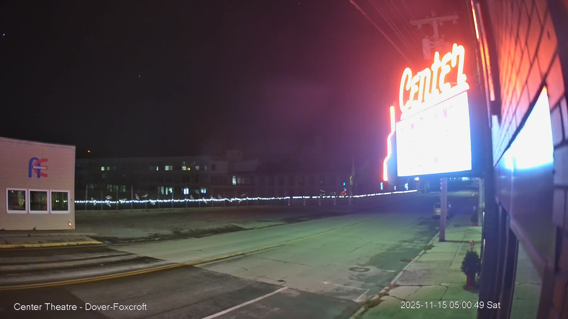 A clear nighttime view of a street shows a bright red and white neon "Center" sign illuminating the right side, with a road, sidewalk, a building featuring a blue logo on the left, and distant string lights beneath a few visible stars.
