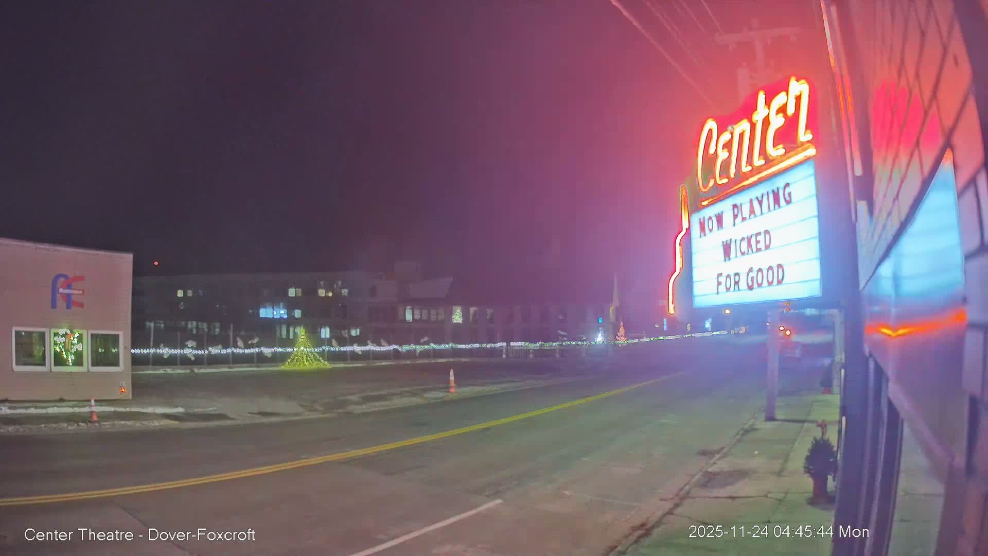 A clear night with visible atmospheric light diffusion illuminates an urban street featuring a bright neon sign on the right, a building with a logo on the left, and distant buildings strung with festive lights including a glowing conical tree across the street.