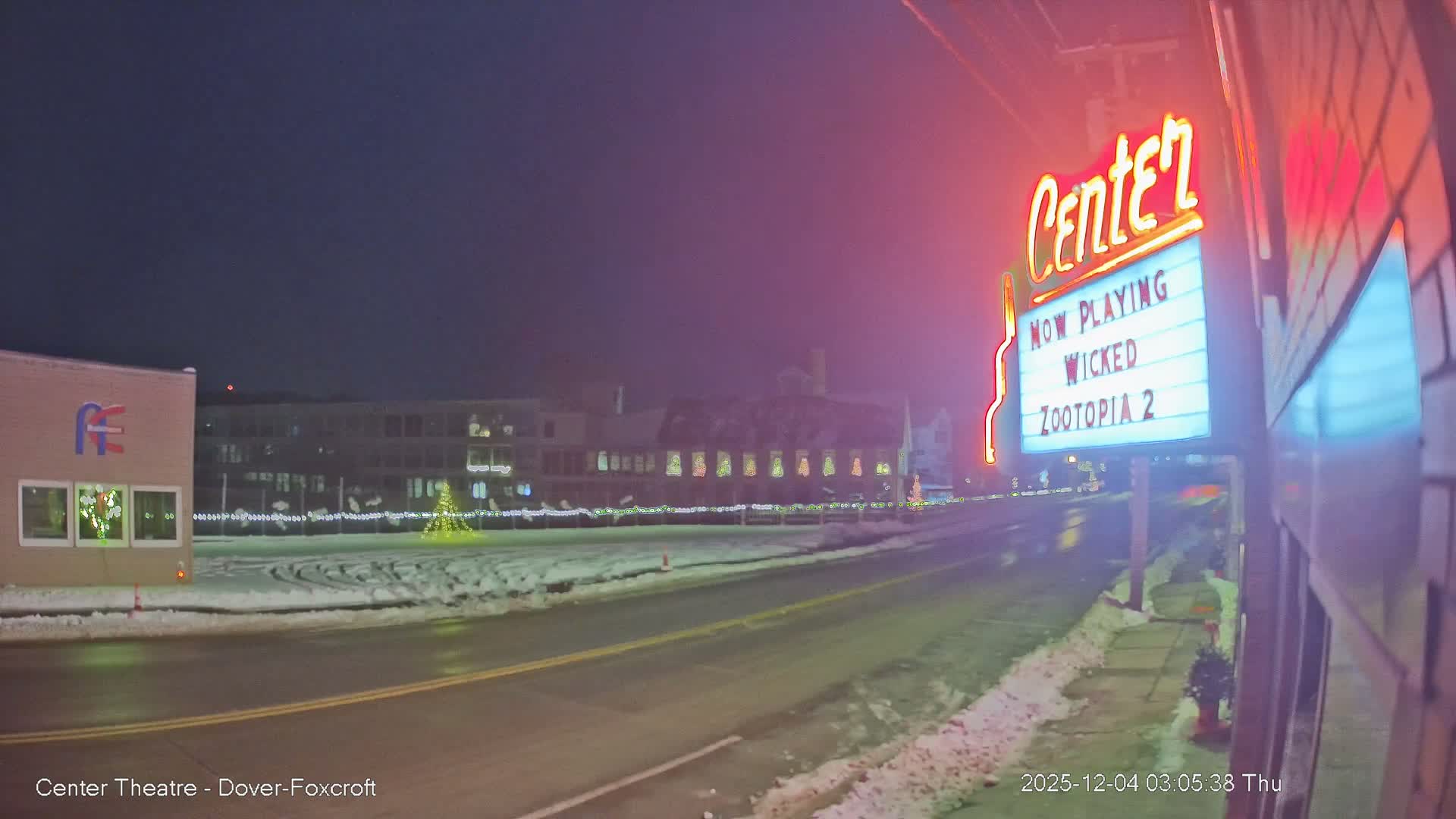 On a clear, cold winter night, a brightly lit movie theater marquee stands beside a snow-lined two-lane road, facing other buildings decorated with string lights and a festive Christmas tree.