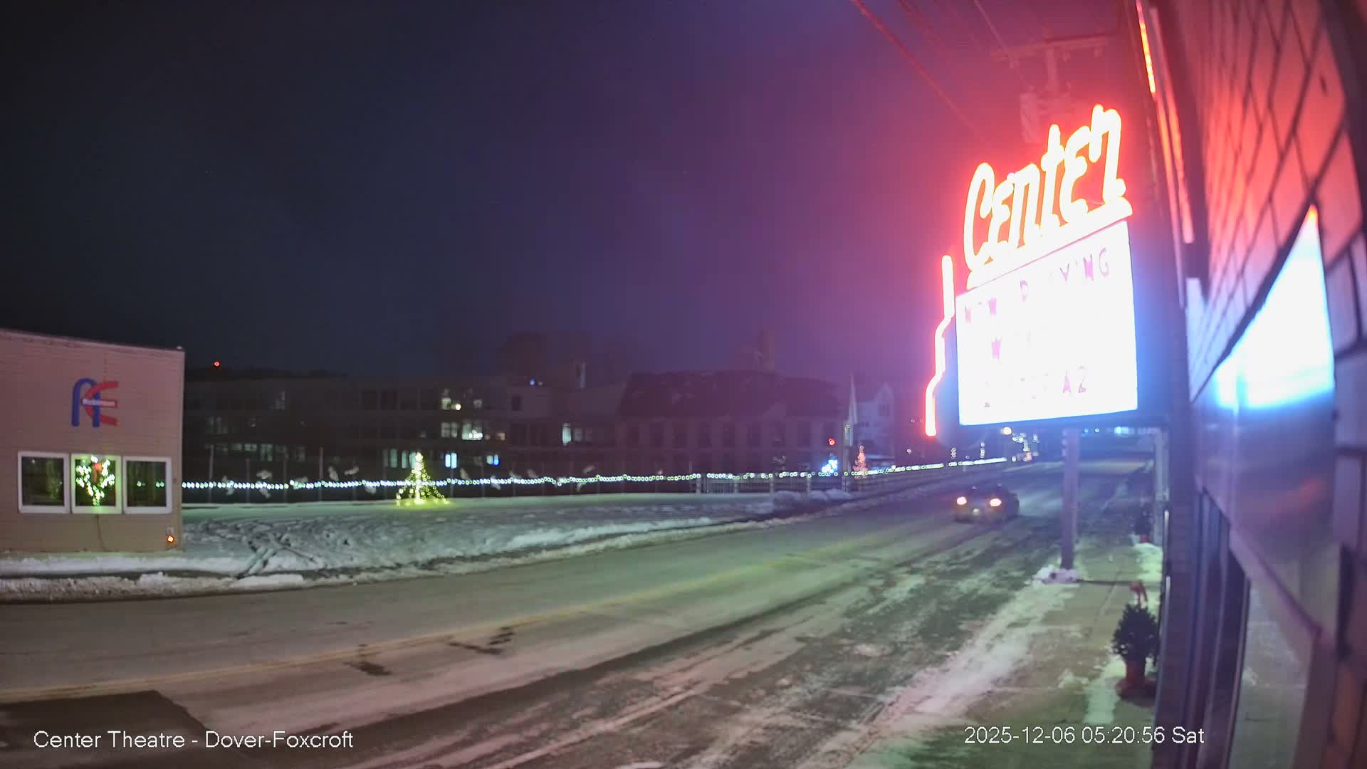A cold, snowy winter night scene shows a brightly lit red neon sign on the right, overlooking a street with a car driving away and background buildings adorned with Christmas lights.