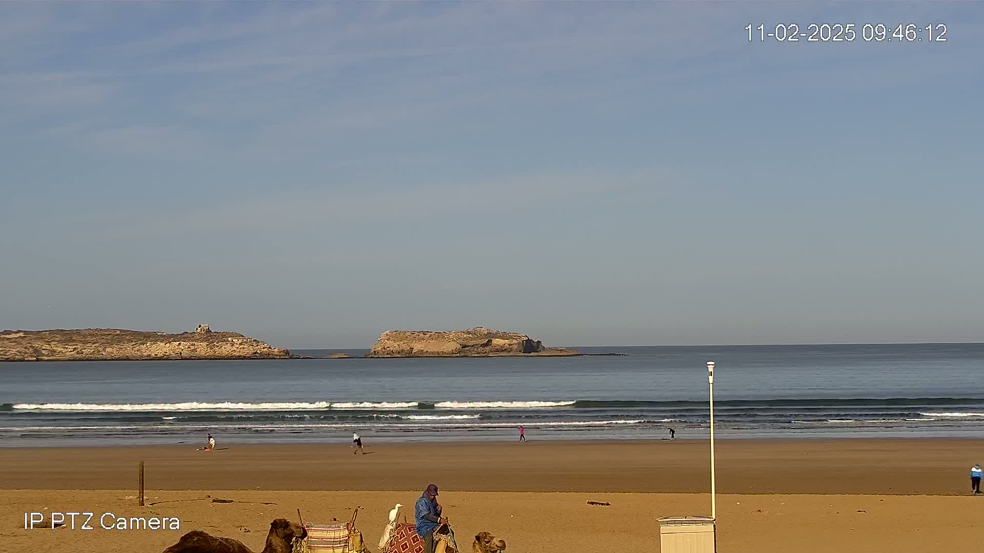 A sandy beach with a few people and camels, gentle waves rolling onto the shore under a mostly clear sky, with two small islands visible in the distance.