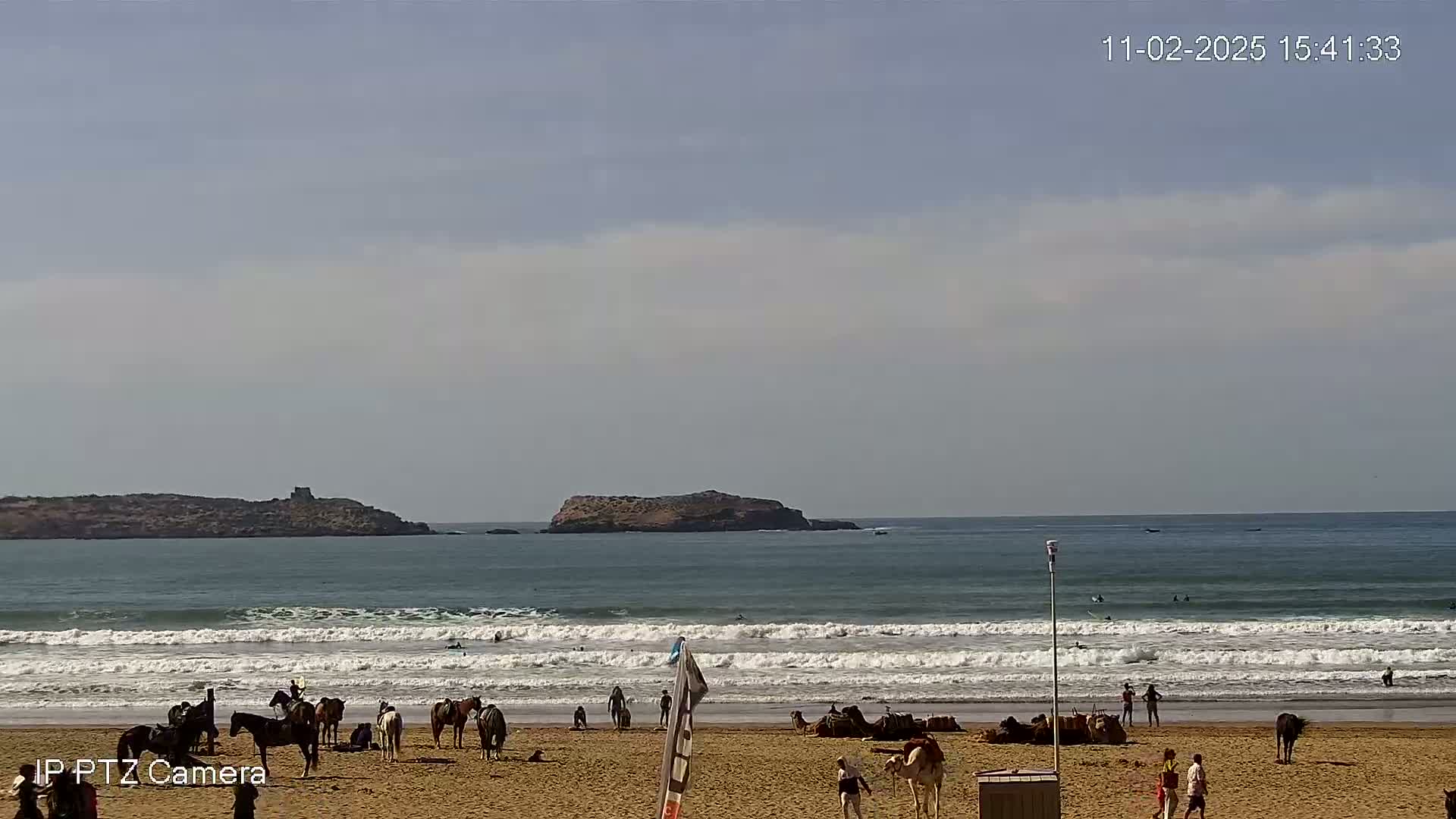 A sandy beach with people, horses, and camels on it, faces a wavy ocean with small islands in the distance under a partly cloudy sky.