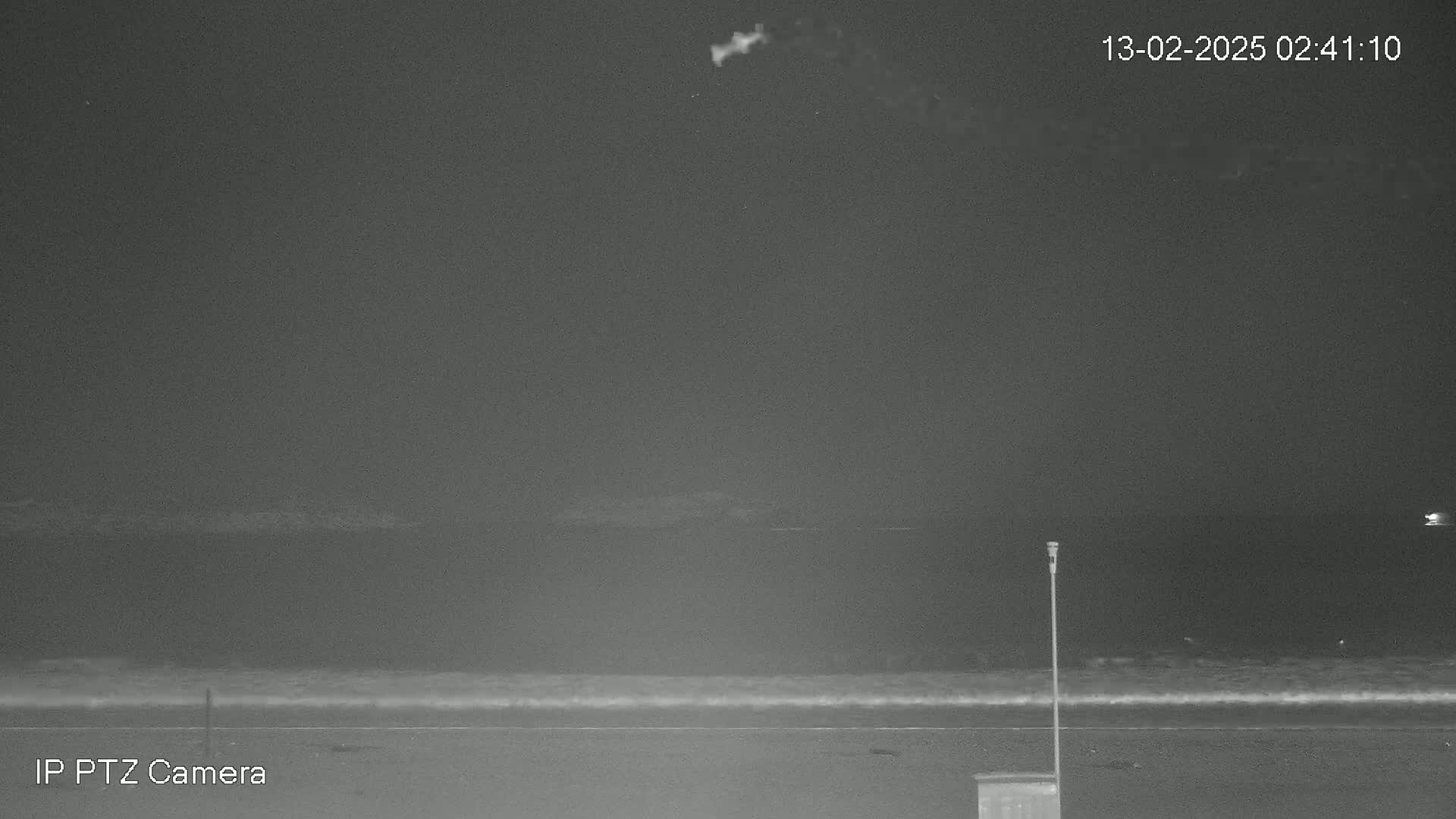 A nighttime, low-light image shows a dark ocean with small waves lapping a shore under a mostly clear sky.