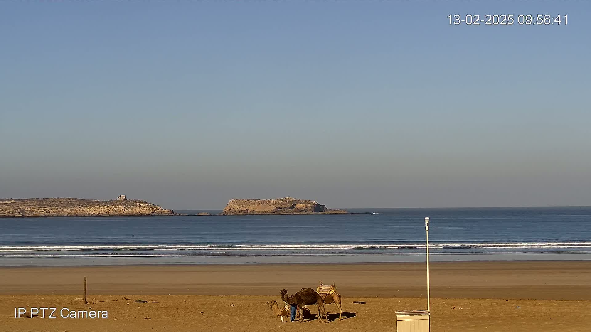 A sandy beach with a camel and two people near a small structure, overlooks a calm ocean with two small islands in the distance under a clear sky.