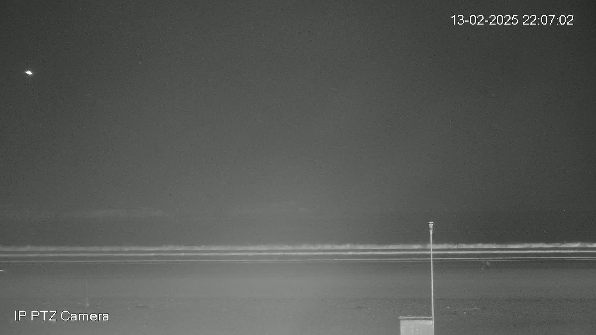 A nighttime beach scene shows a dark sky with a single bright light, calm waves lapping the shore, and a single pole standing near a small structure.