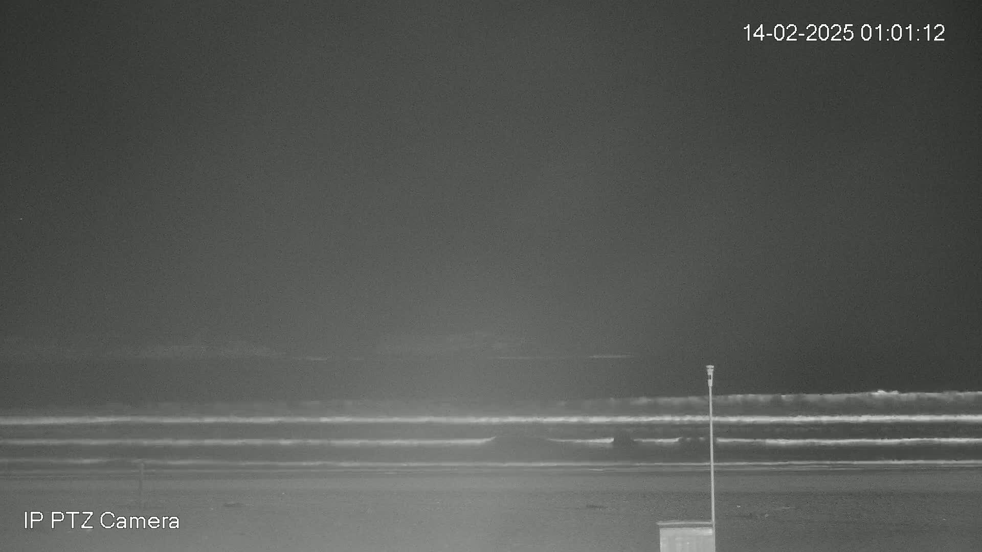 A nighttime, low-light view shows a dark beach with calm waves gently rolling onto the shore under a mostly clear sky.