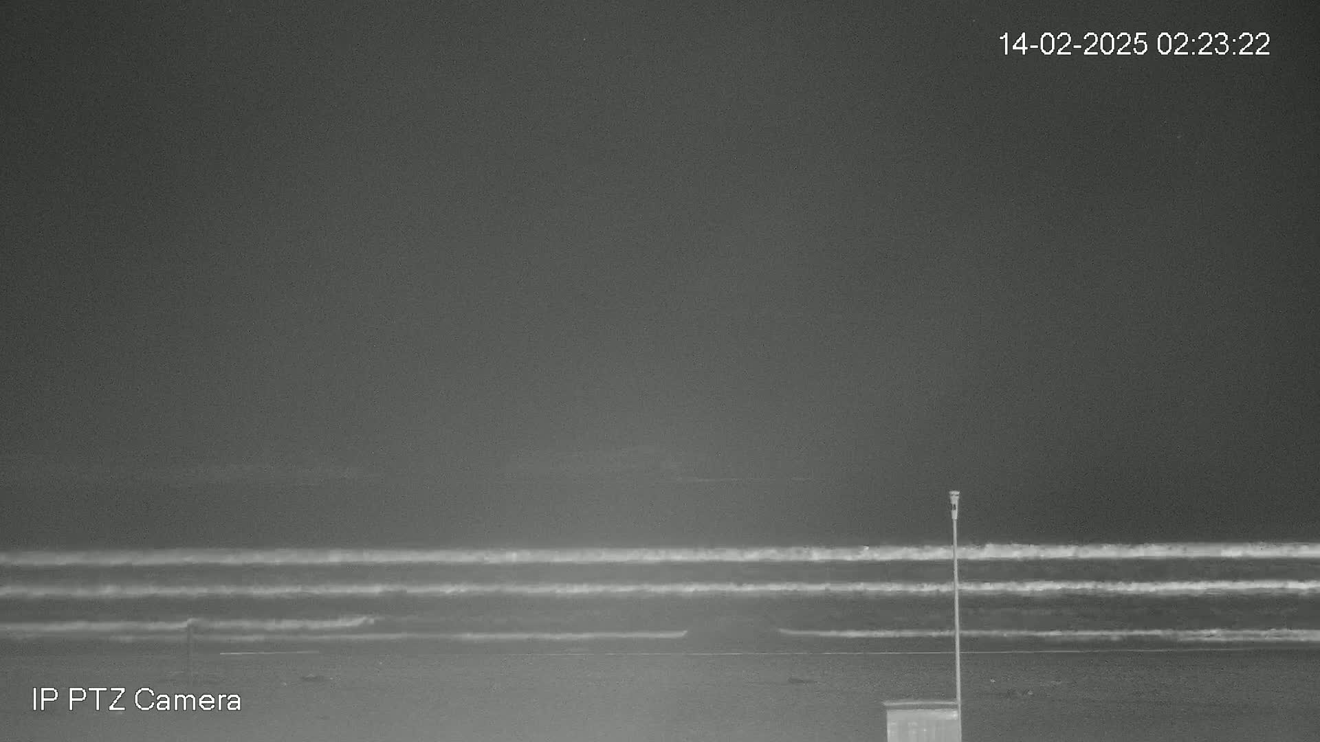 A nighttime, low-light grayscale image shows a dark beach with a calm ocean featuring three parallel lines of small waves under a dark sky.