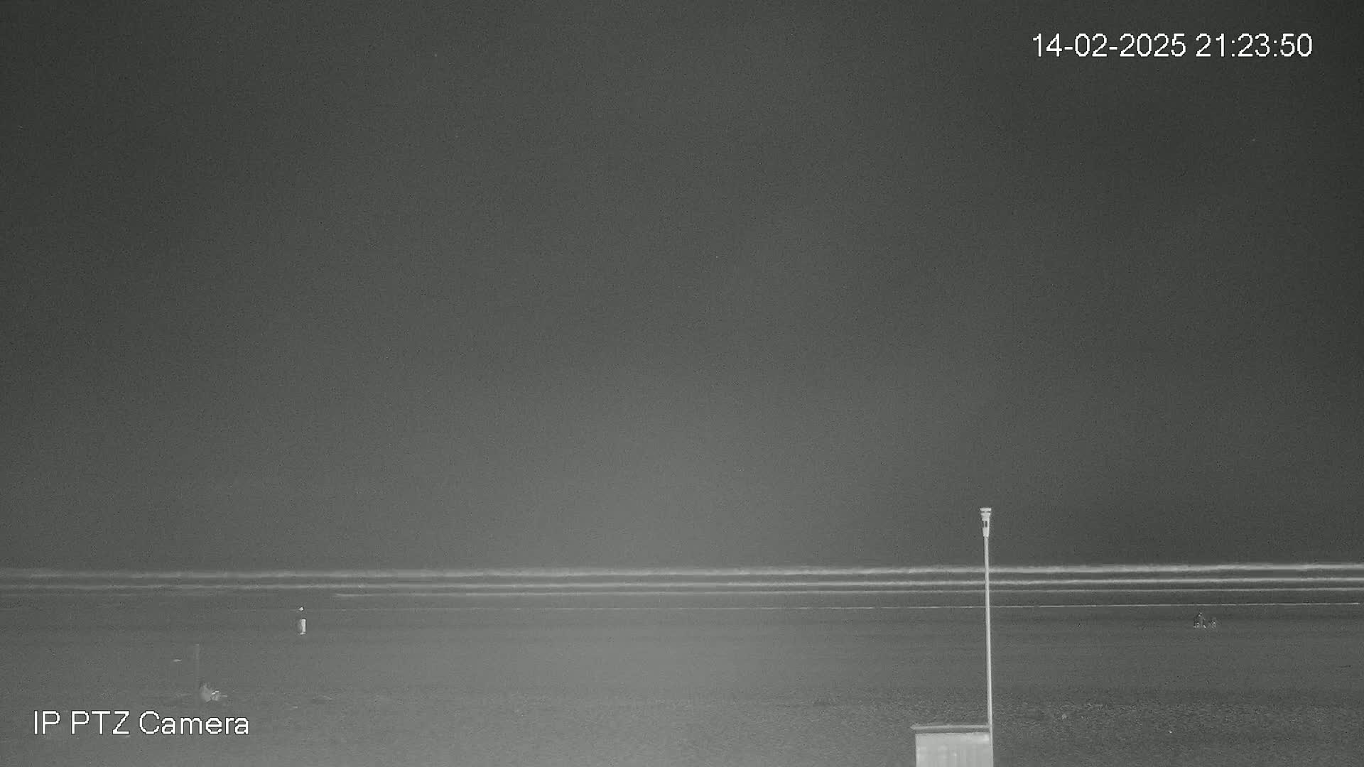 A nighttime, low-light view of a dark beach with a calm ocean and a single pole standing near a small structure.