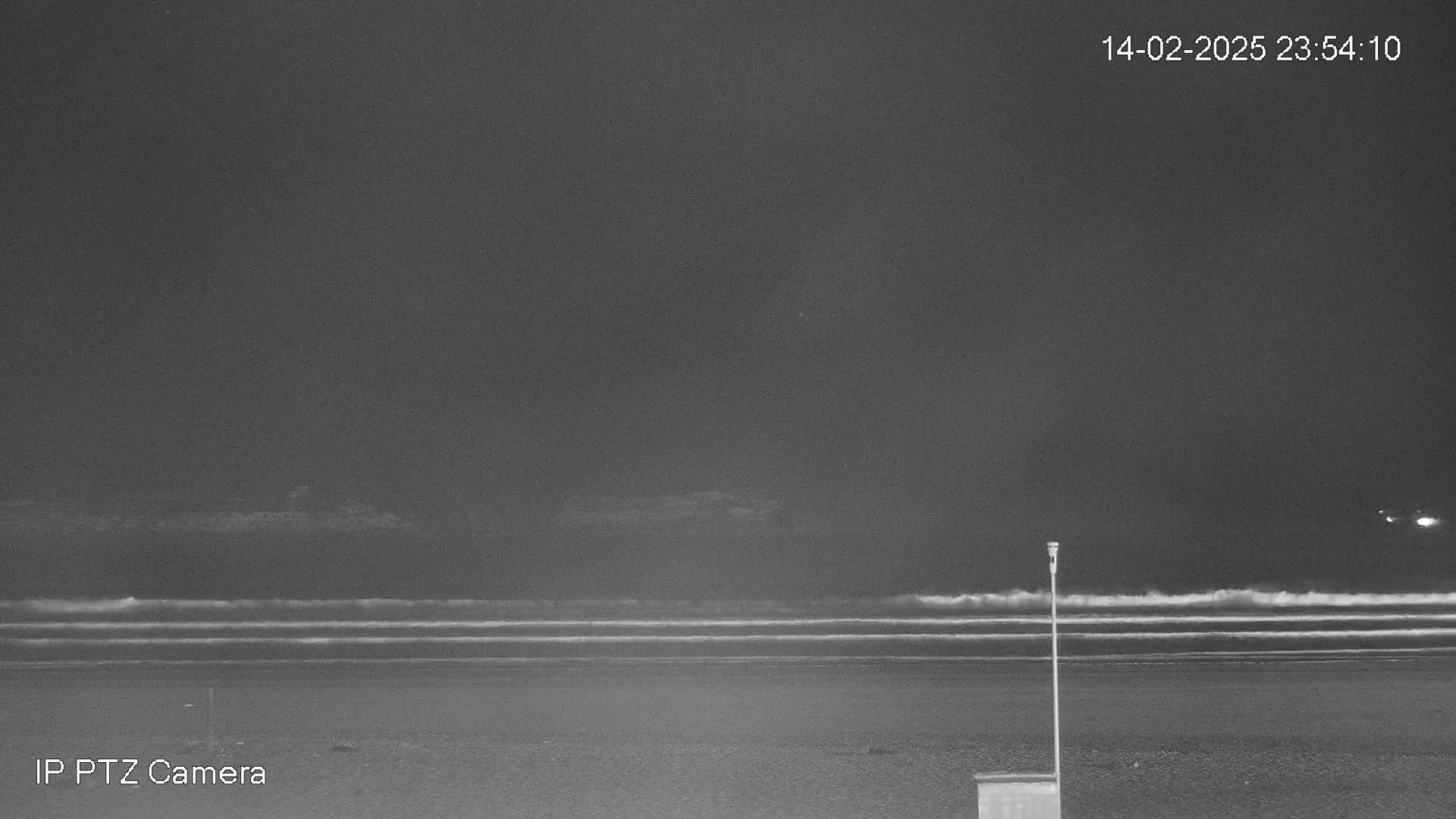 A dark nighttime beach scene shows gently rolling waves under a clear, dark sky, with a single post and a small structure visible on the sand.