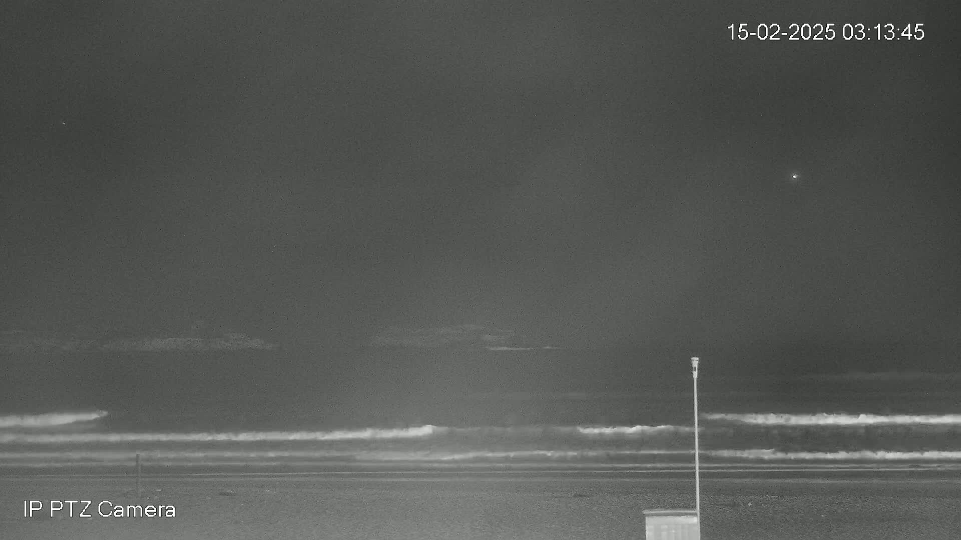 A nighttime, grayscale image shows a dark ocean with small waves lapping a beach under a cloudy sky.