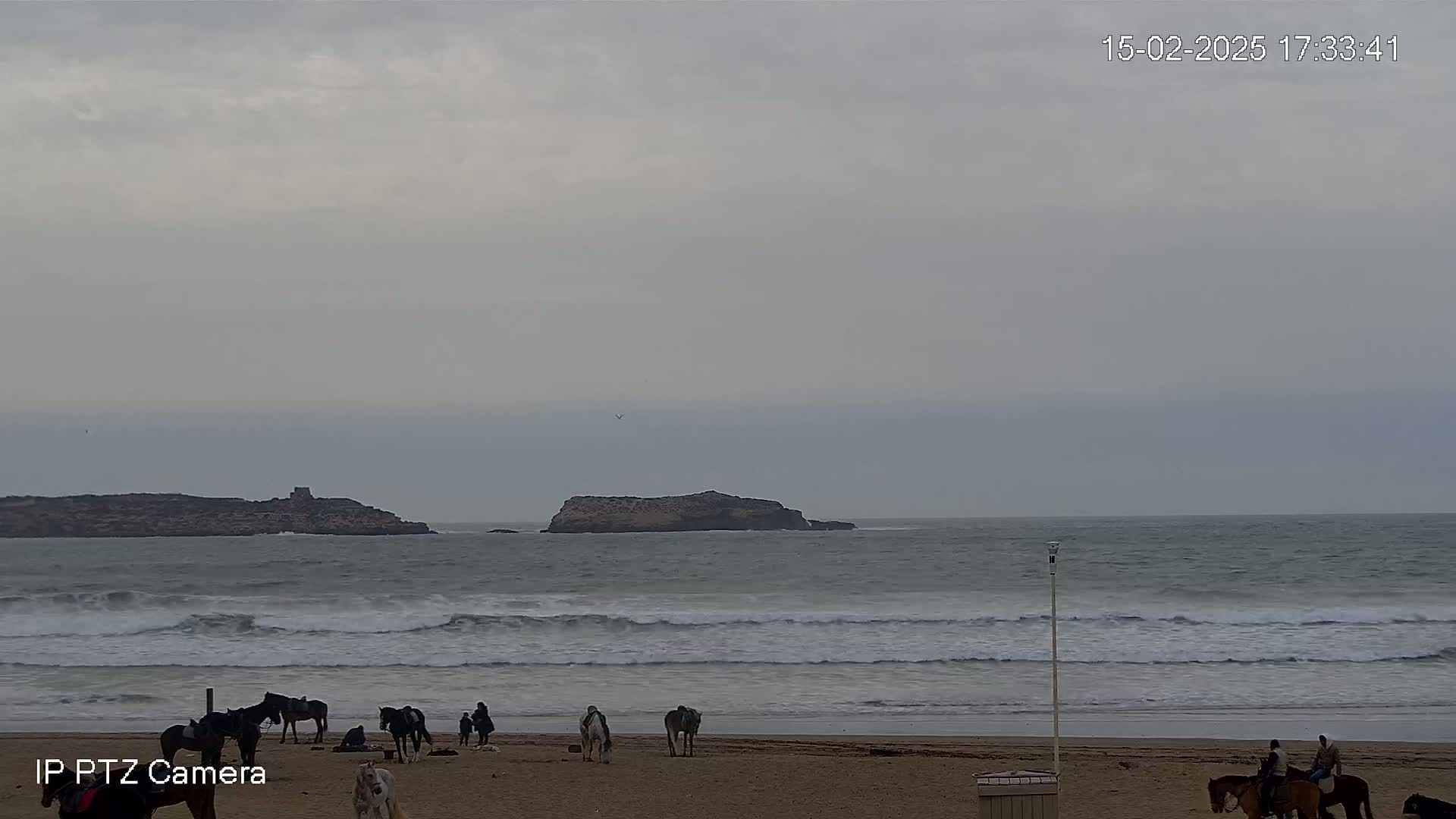 A mostly cloudy day shows several horses and riders on a sandy beach near a rocky island in the ocean with moderate waves.